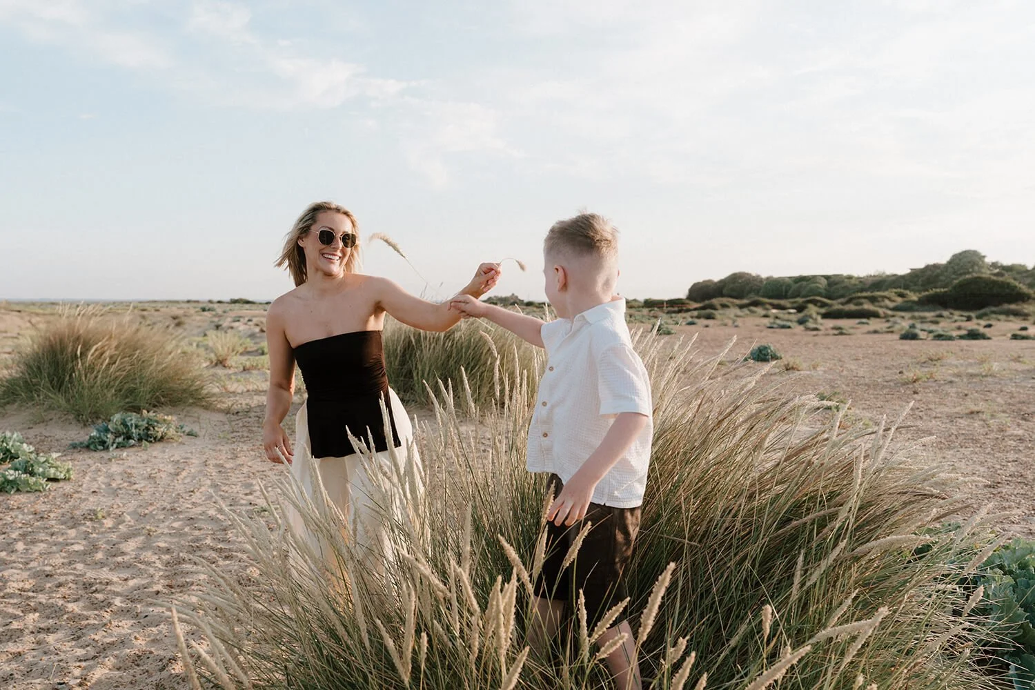 A woman in sunglasses and a strapless black top playing with a young boy in a white shirt among tall grasses in a sandy desert landscape with scattered greenery.