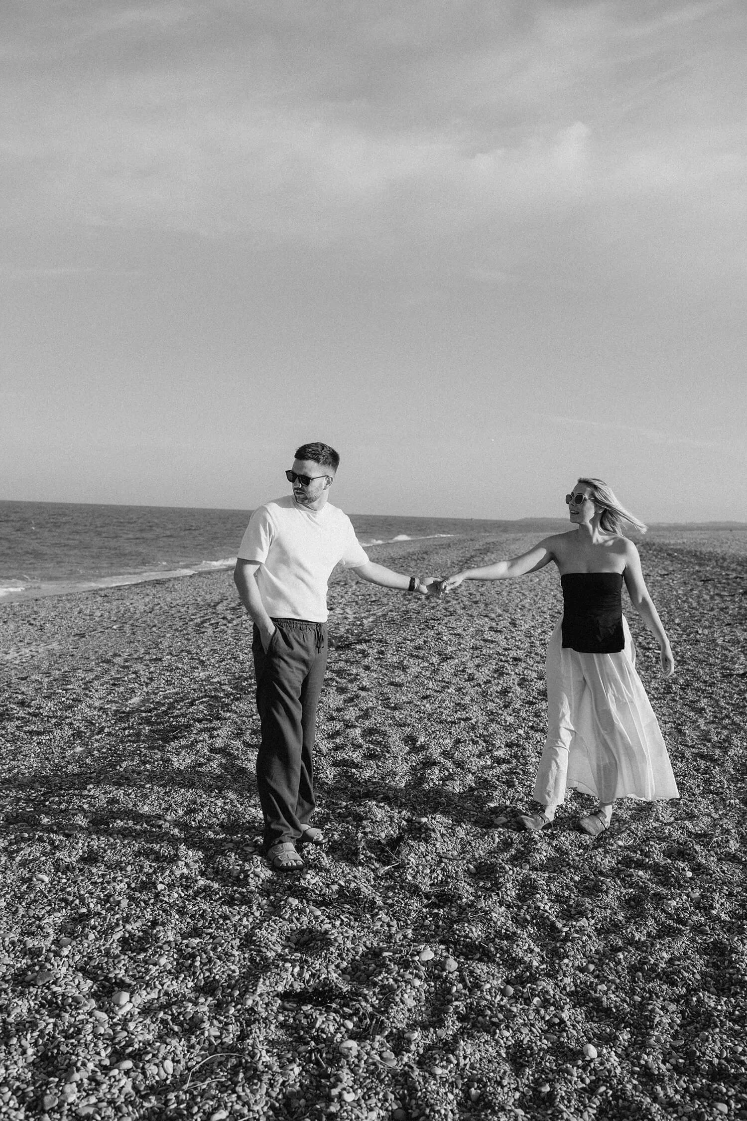 A black-and-white photo of a man and woman holding hands while walking on a pebble beach near the ocean, with the woman smiling and the man looking away, both wearing sunglasses.