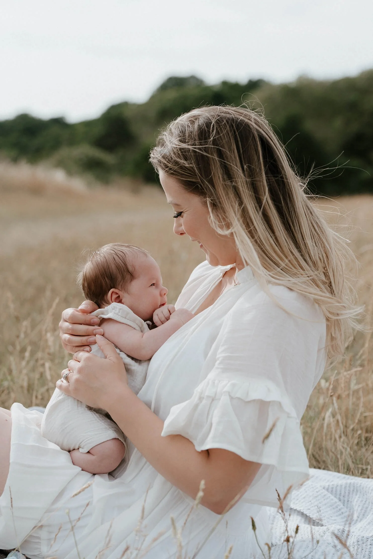 A woman holding a baby in an outdoor field with grass and trees in the background.