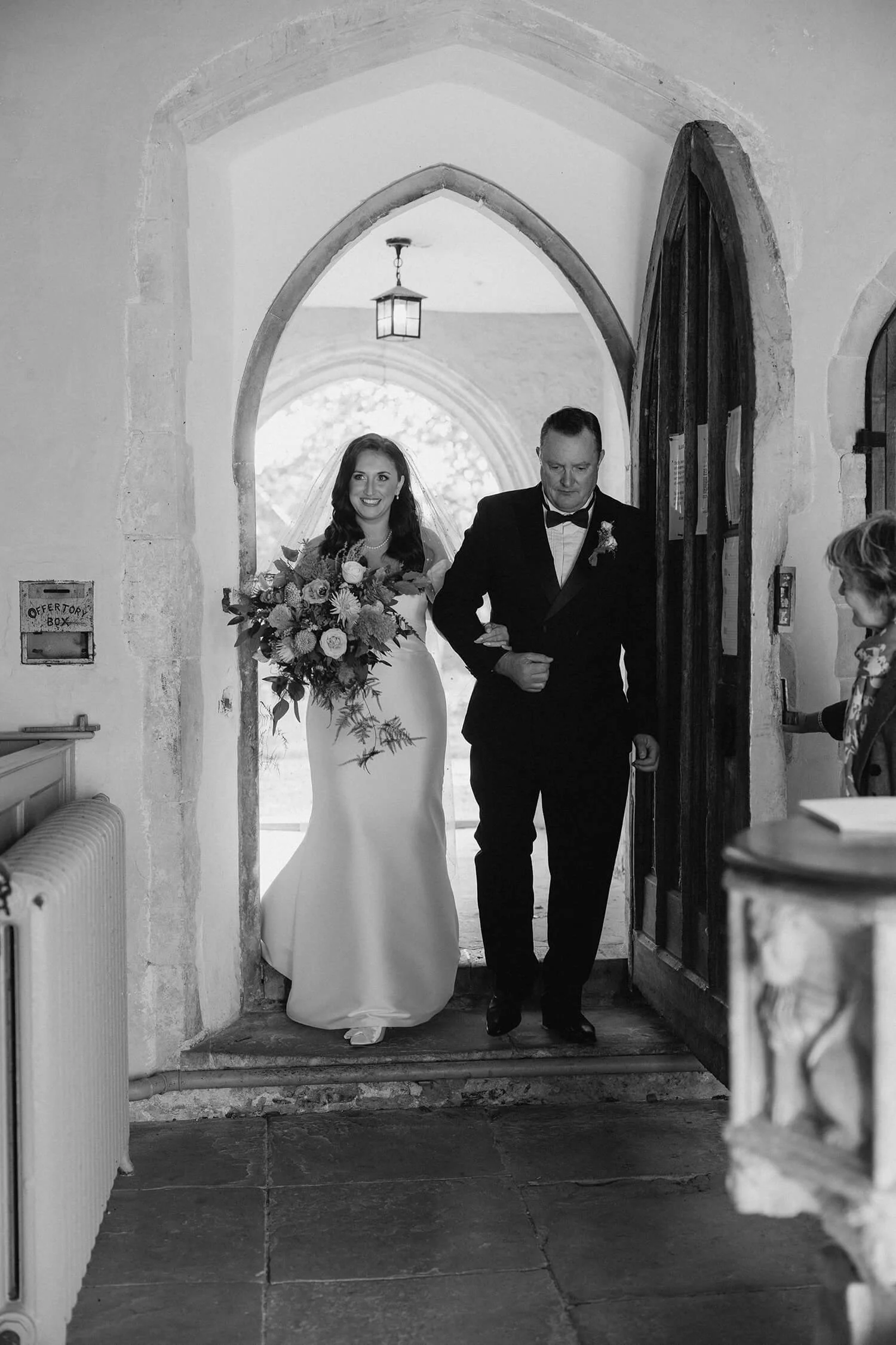 A bride in a white wedding dress holding a bouquet, walking out of a church with a man in a tuxedo, possibly her father, during a wedding ceremony.