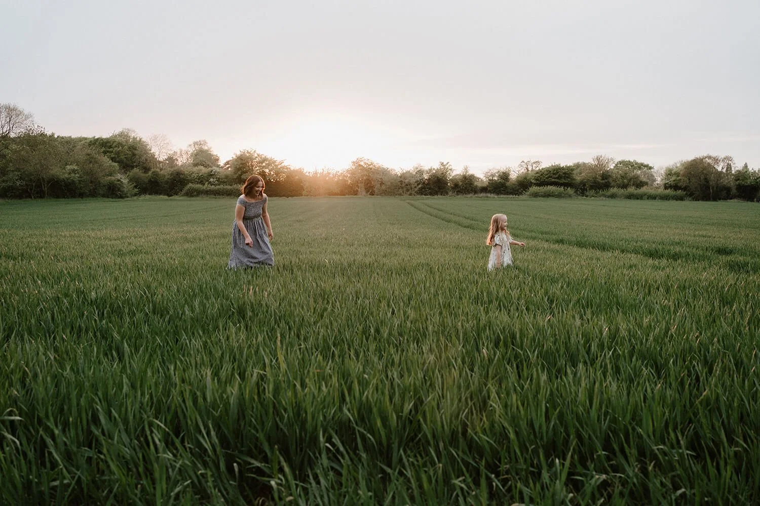A woman and a young girl walking through a green field at sunset.