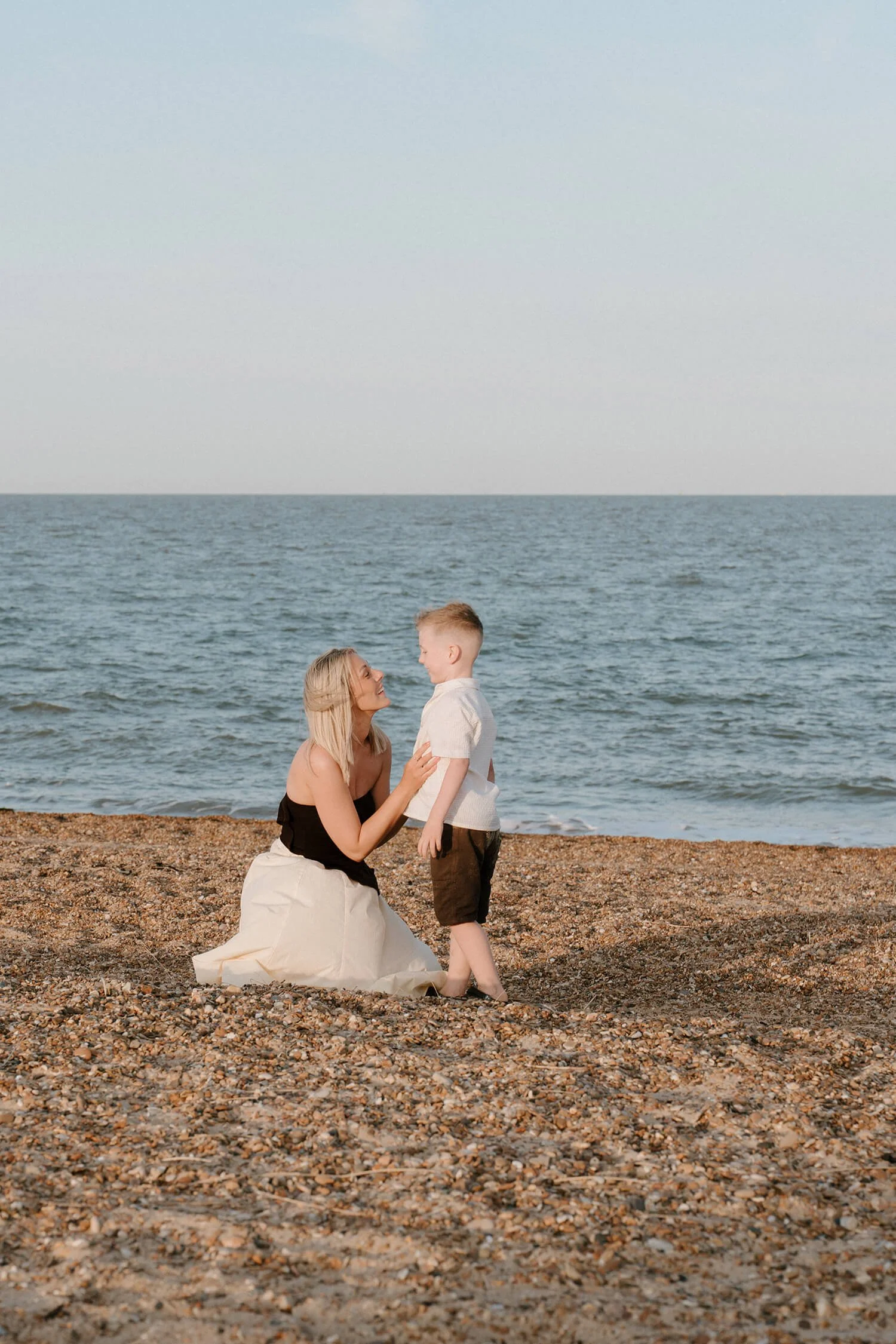 A woman kneeling on a sandy beach holding a young boy's arm, with the ocean in the background during the daytime.