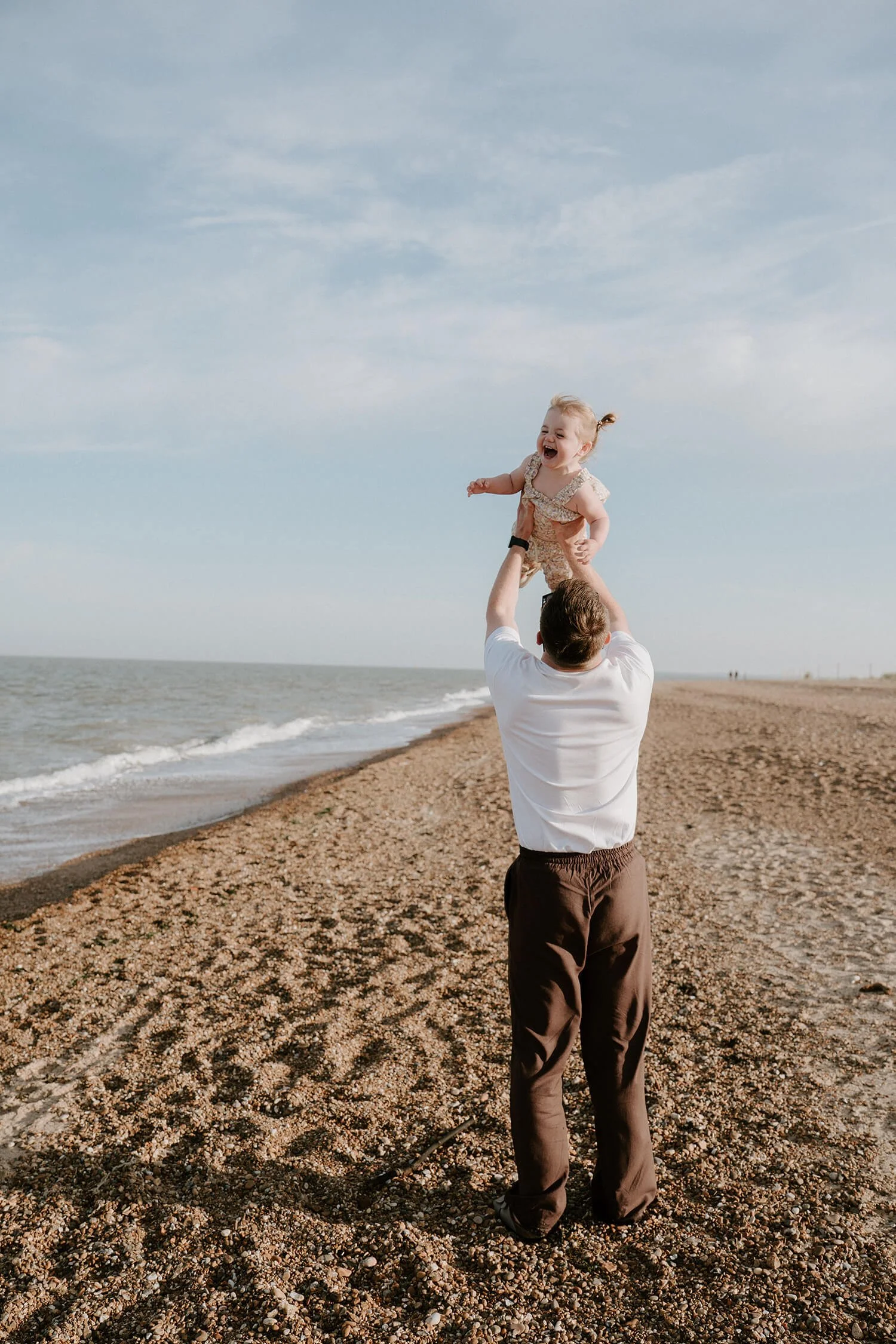 A man holding a young girl in the air on a beach, with the ocean and sky in the background, both smiling and appearing happy.