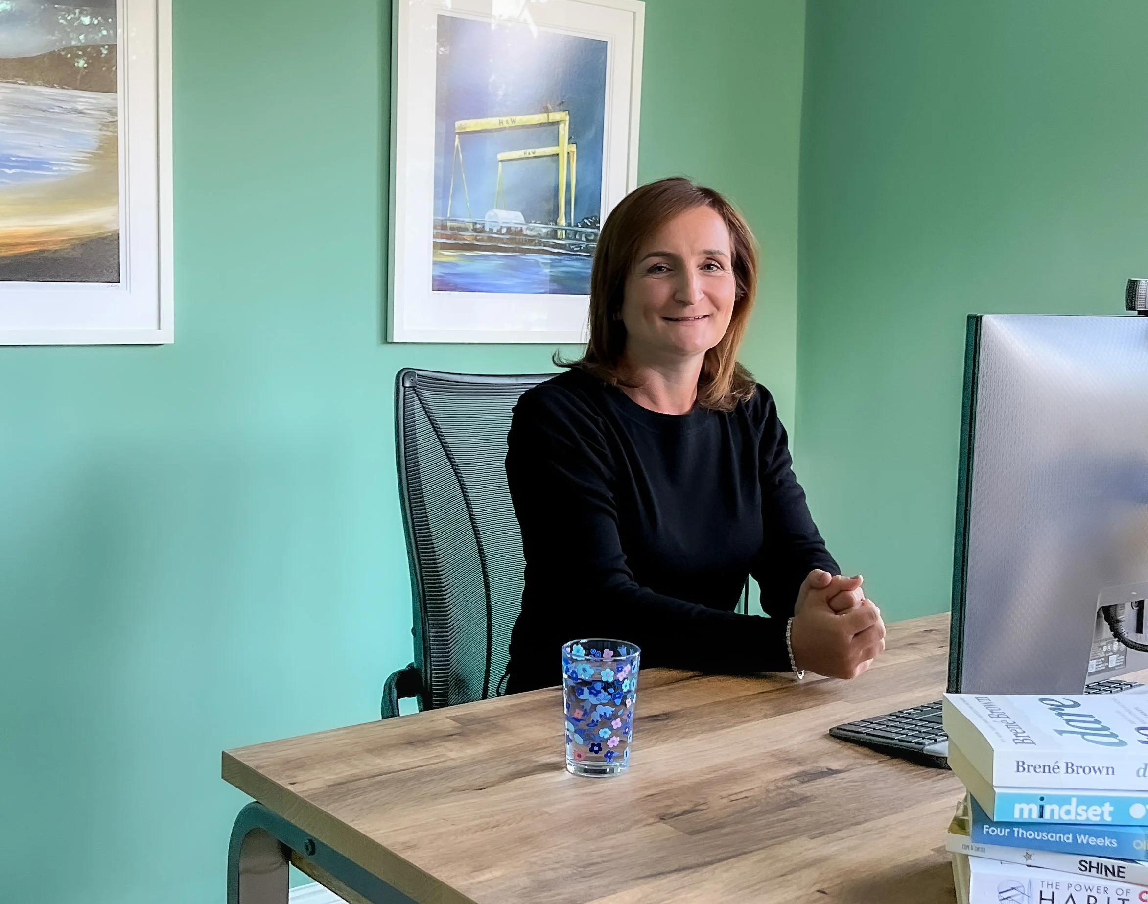 A woman with shoulder-length brown hair wearing a black long sleeve shirt sitting at a wooden desk in an office, smiling, with a computer monitor, a book pile, and a patterned blue glass of water on the desk, with green walls and framed artwork in the background.