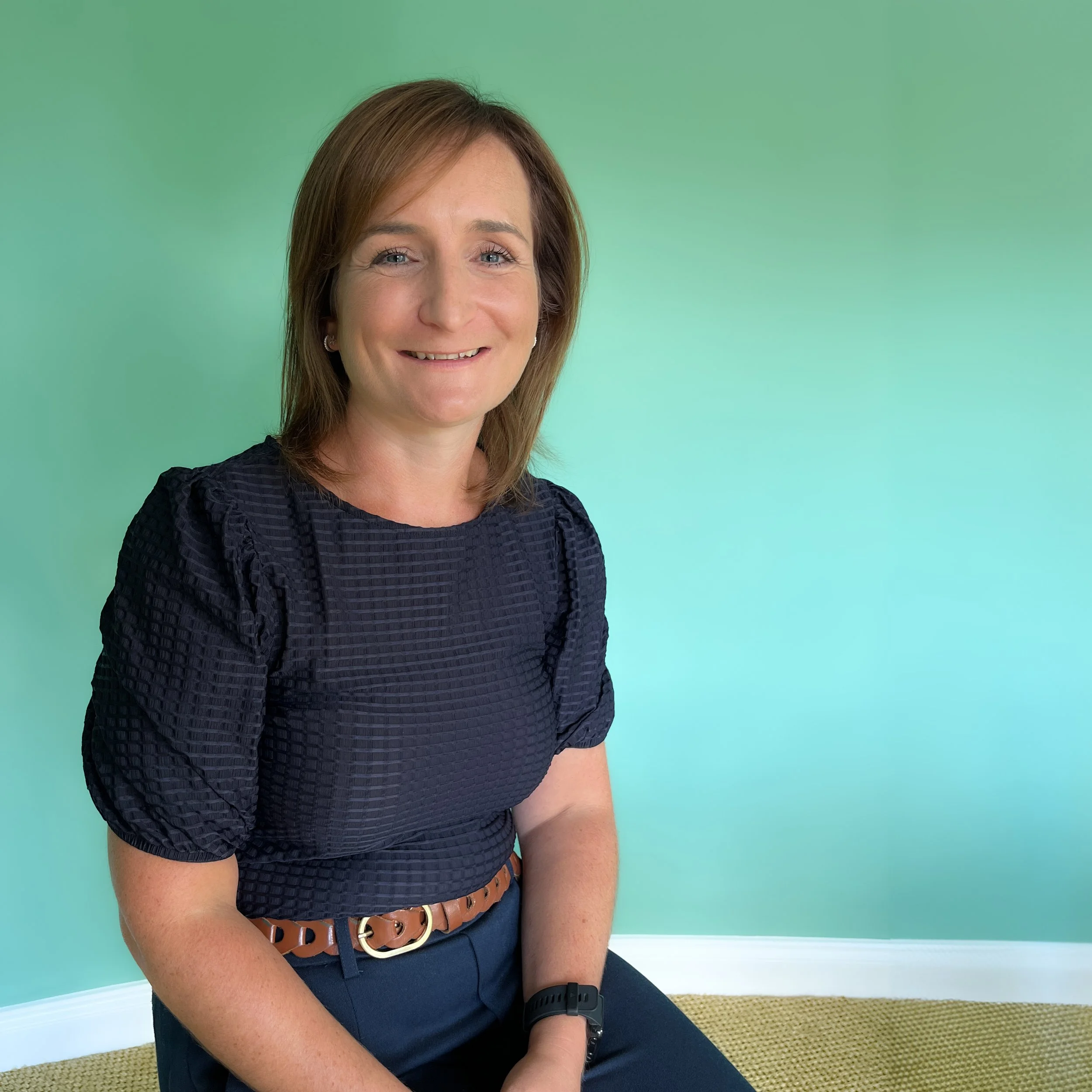 A woman with brown hair wearing a navy textured blouse, sitting on a yellow carpet, with a light green wall in the background, smiling at the camera.