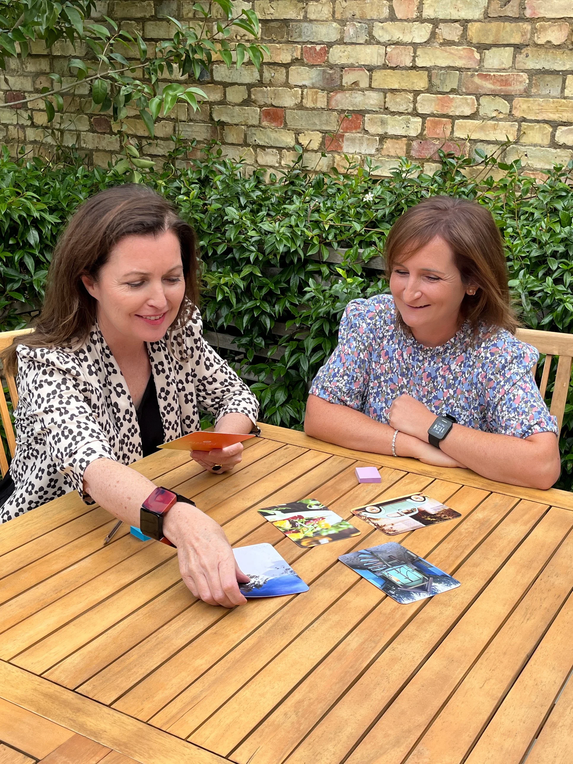 Two women sit at a wooden table outdoors, playing a card game. They are smiling and appear to be enjoying themselves, with a brick wall and green bushes in the background.