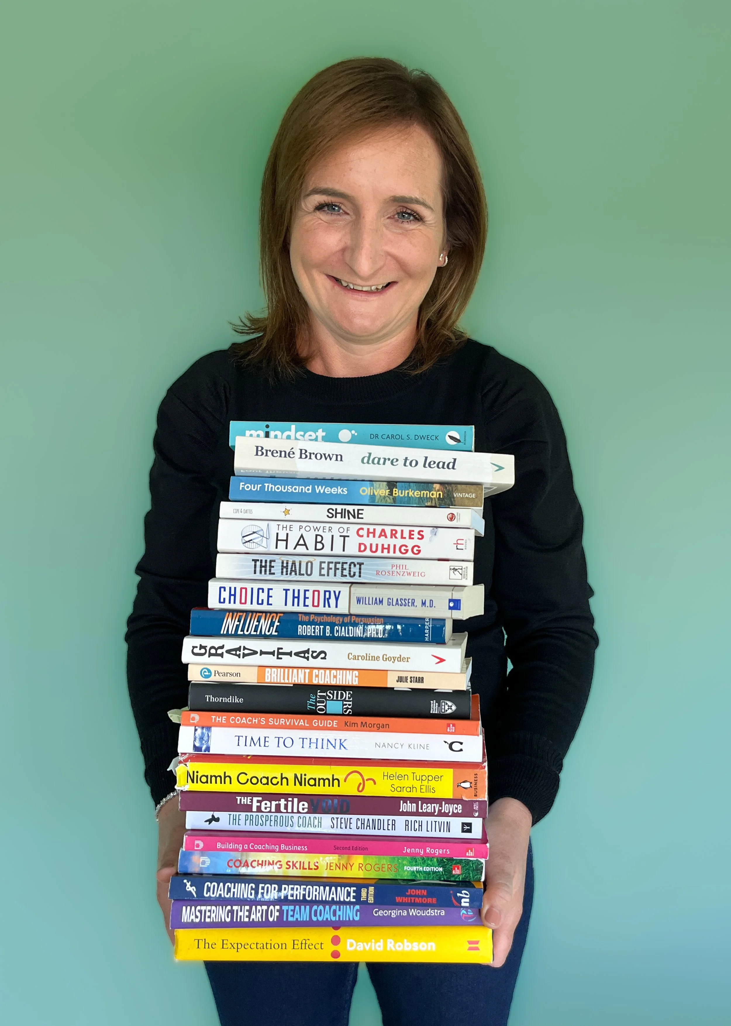 A woman with shoulder-length brown hair smiling and holding a stack of books about coaching, leadership, and psychology against a green wall.