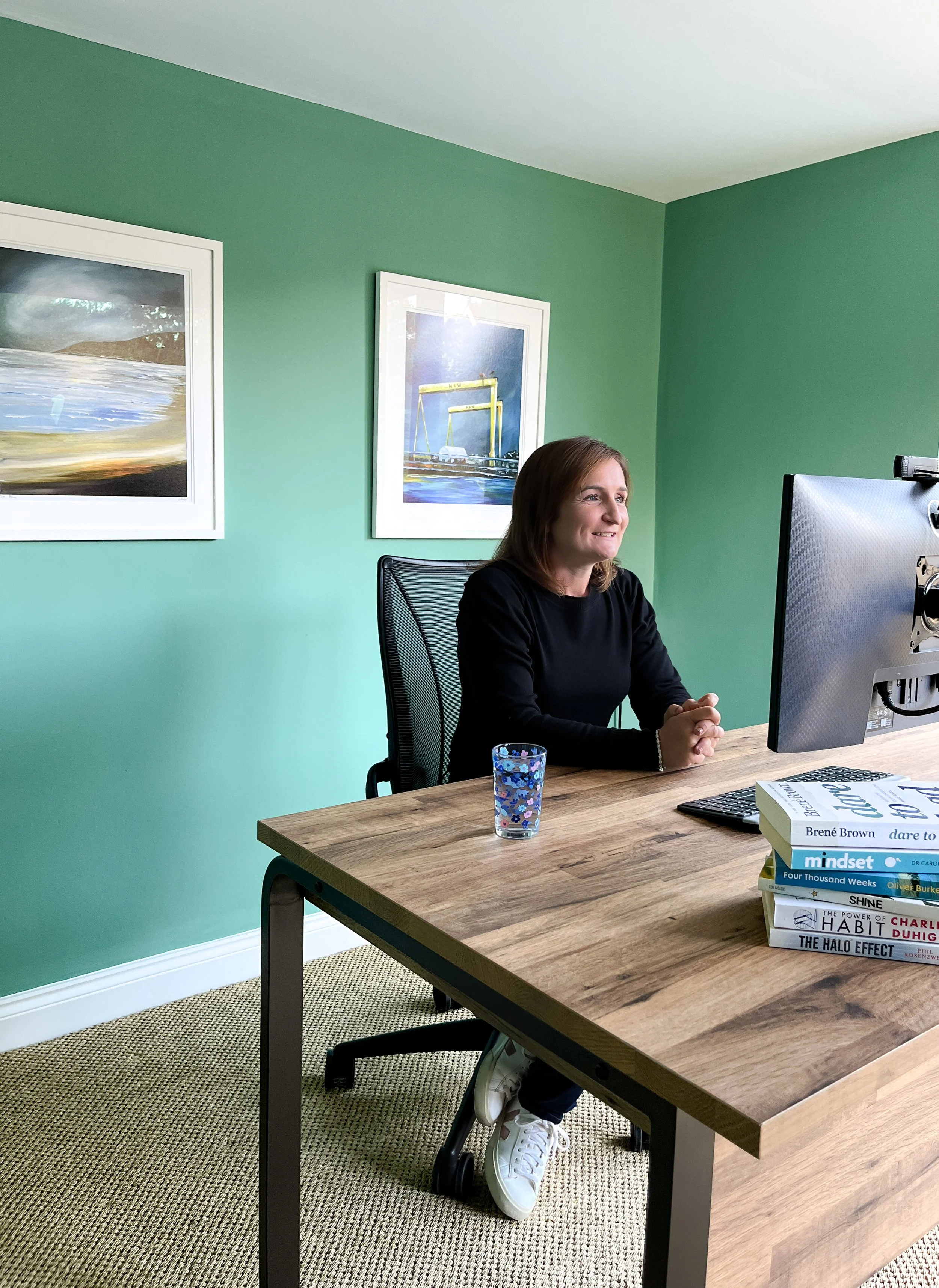A woman sits at a wooden desk in a green-walled office, smiling and looking at a computer monitor. There are books stacked on the desk and framed artwork on the wall behind her.