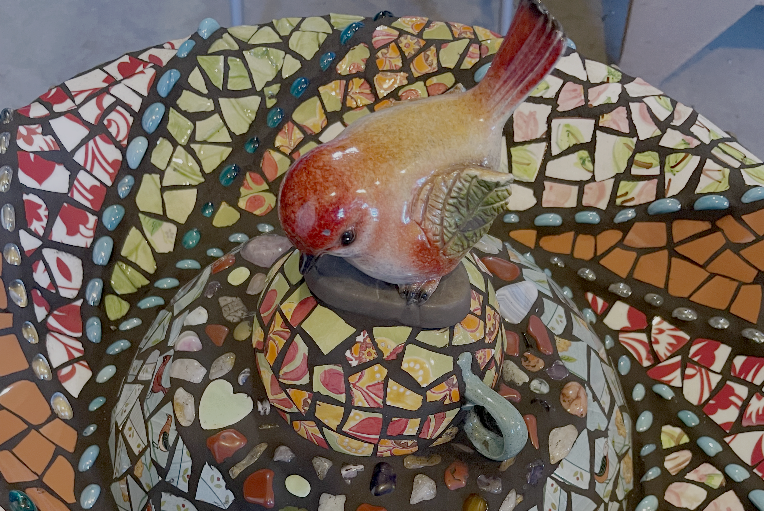 Decorative mosaic table with a colorful mosaic pattern, a ceramic bird figurine with red, orange, and yellow hues, and a small ceramic teapot.