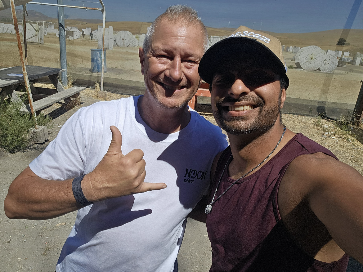 Two men smiling: one giving a thumbs-up sign, both taking a selfie outdoors with a shooting range in the background, featuring targets and rolling hills.