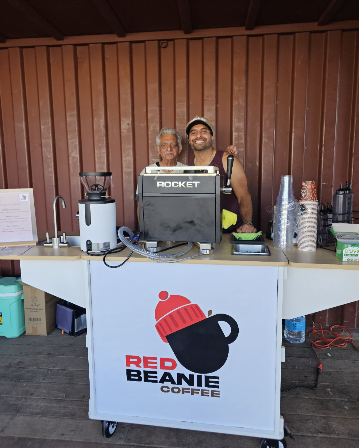 Two men behind a coffee counter with a "Red Beanie Coffee" logo, one smiling broadly, both standing in front of a brown wooden wall.