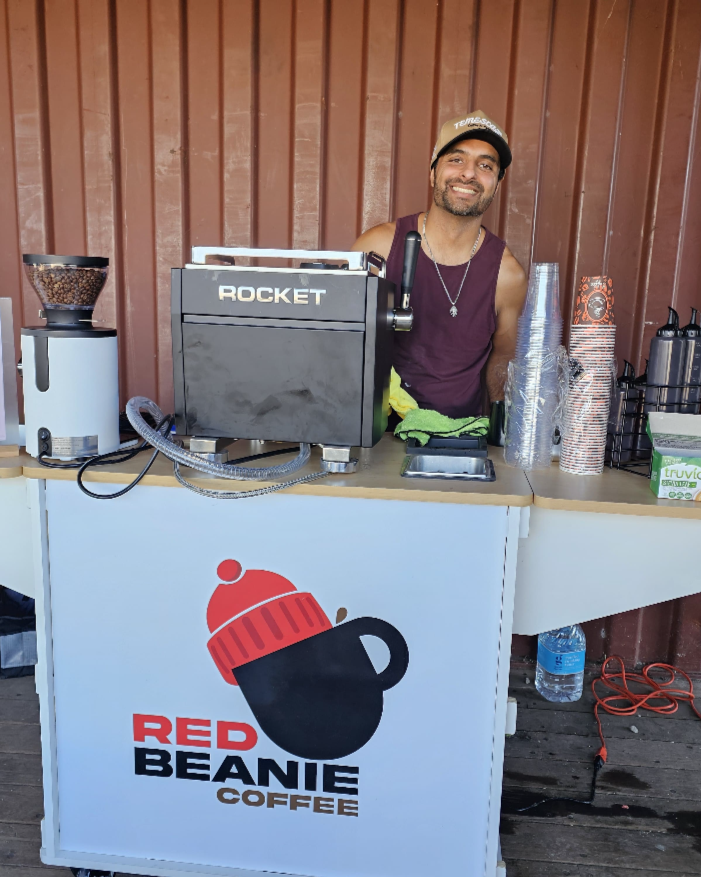 A man standing behind a coffee stand labeled "Red Beanie Coffee" with a logo of a red beanie hat over a black coffee mug. He is smiling, wearing a maroon tank top and a beige cap, with cups and coffee-making equipment on the counter.