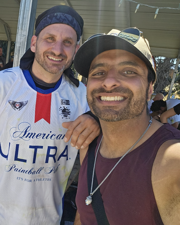 Two men smiling for a selfie outdoors, one wearing a baseball cap and sunglasses, the other wearing a headscarf, with a covered pavilion in the background.