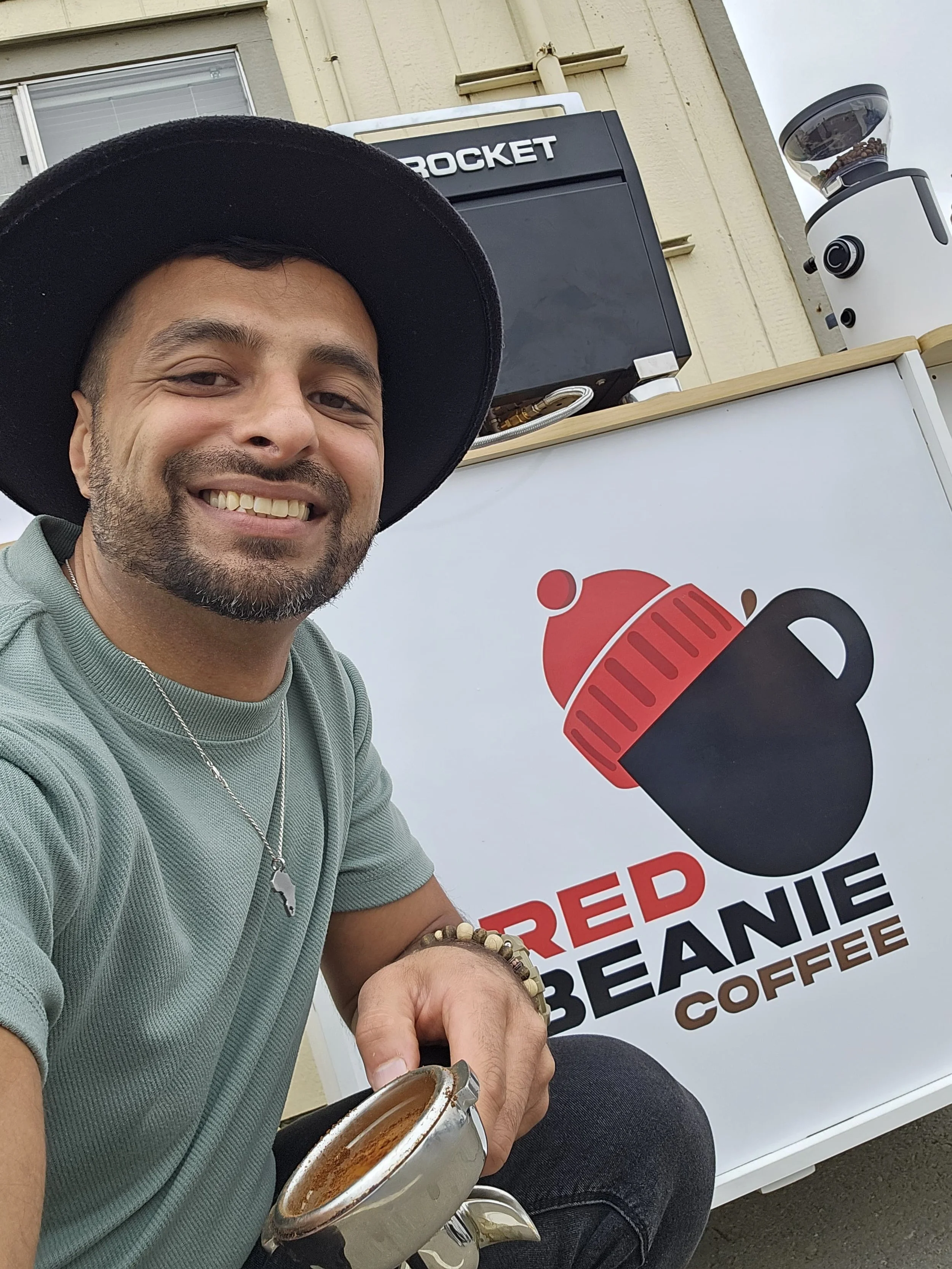A smiling man wearing a black hat and green shirt, holding a cup of coffee, sitting in front of a sign that reads 'Red Beanie Coffee' with a logo of a black mug wearing a red beanie.