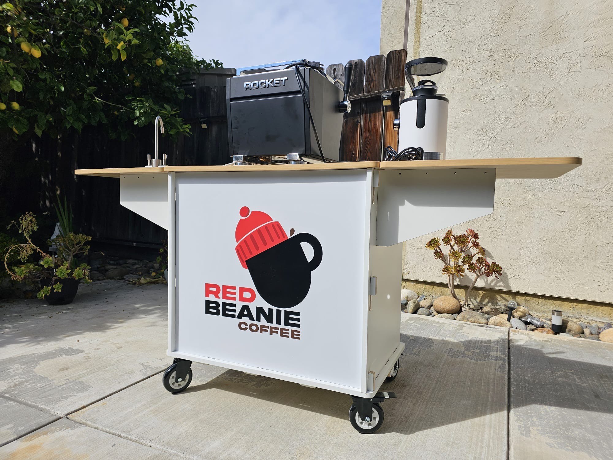 Mobile coffee cart with a logo reading 'Red Beanie Coffee', featuring a black coffee mug wearing a red beanie, on a concrete sidewalk next to a house wall and some plants.
