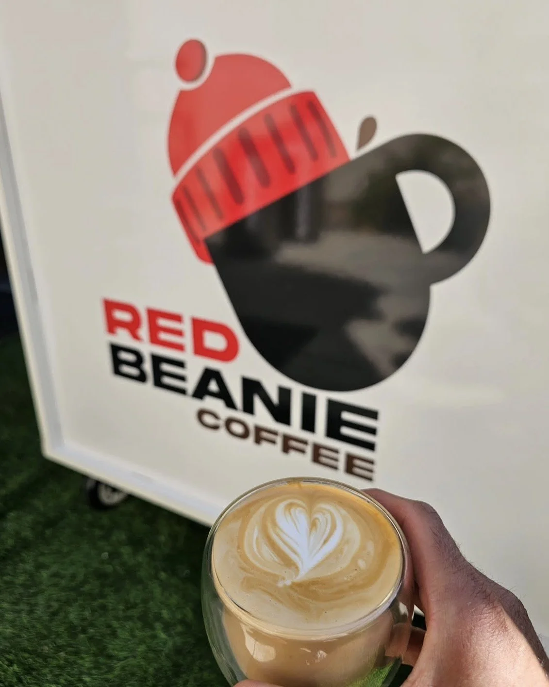 A hand holding a glass of coffee with latte art in front of a sign for Red Beanie Coffee, featuring a black mug with a red beanie on top and the text 'RED BEANIE COFFEE'.