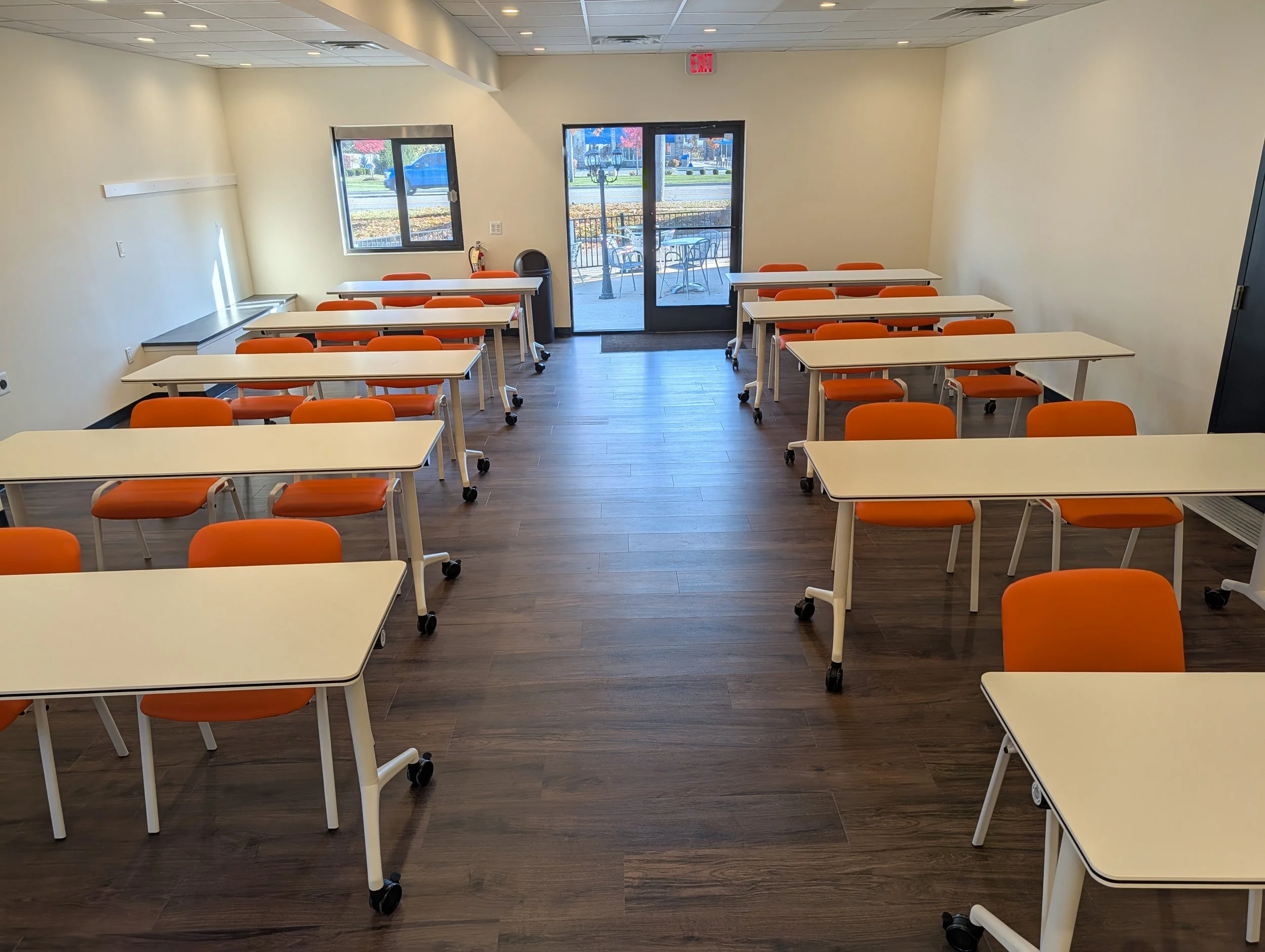 Empty classroom with white desks and orange chairs, facing a door with a glass window, outside patio, and window with trees and red buildings visible.