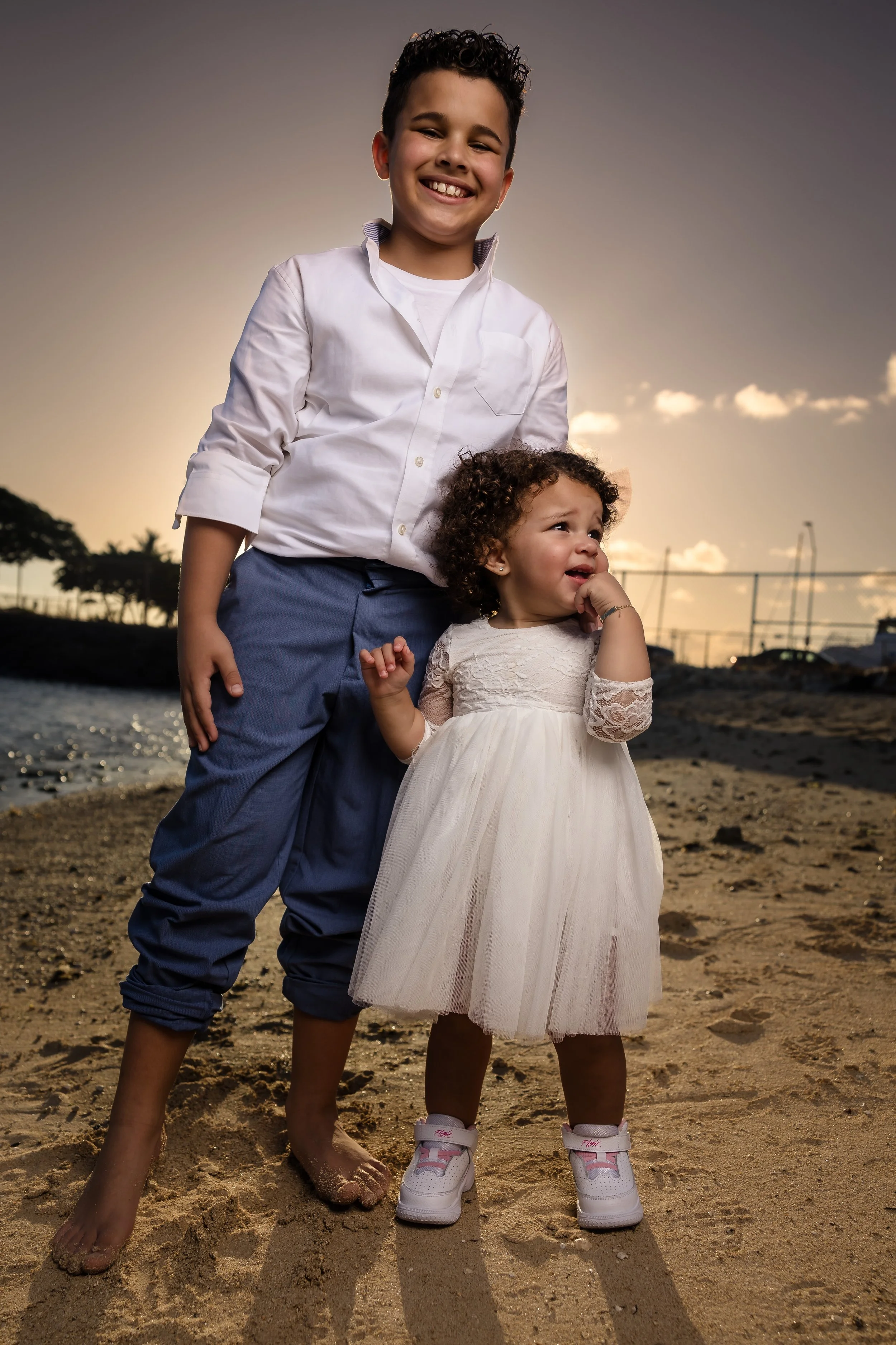 Two children, a boy and a girl, standing on a beach at sunset. The boy is smiling and wearing a white shirt and blue pants, while the girl appears to be upset, wearing a white dress and sneakers. The background shows a fence, water, and a partly clou