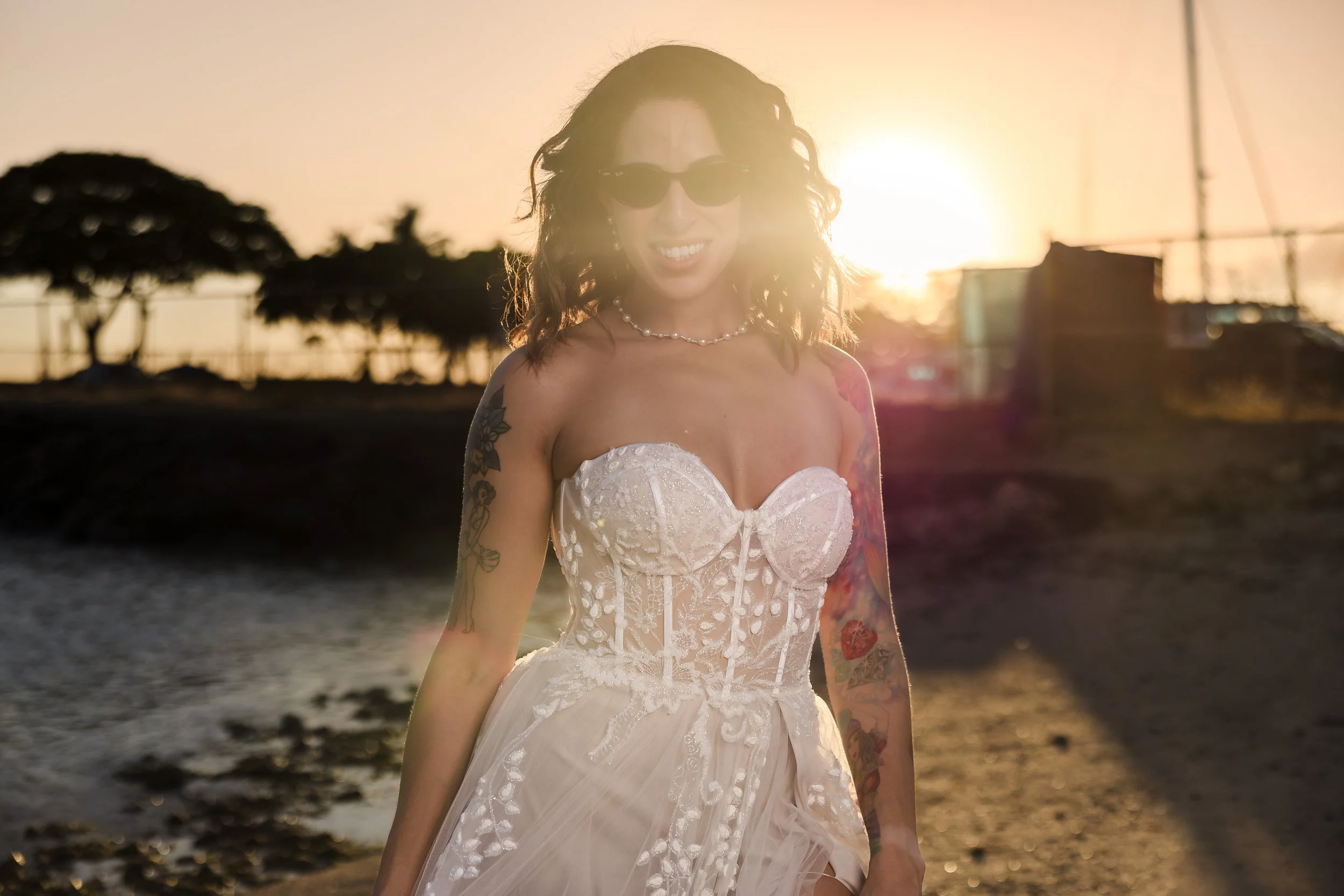 Woman with curly hair wearing sunglasses and a white dress with lace details, standing on a beach at sunset, smiling.