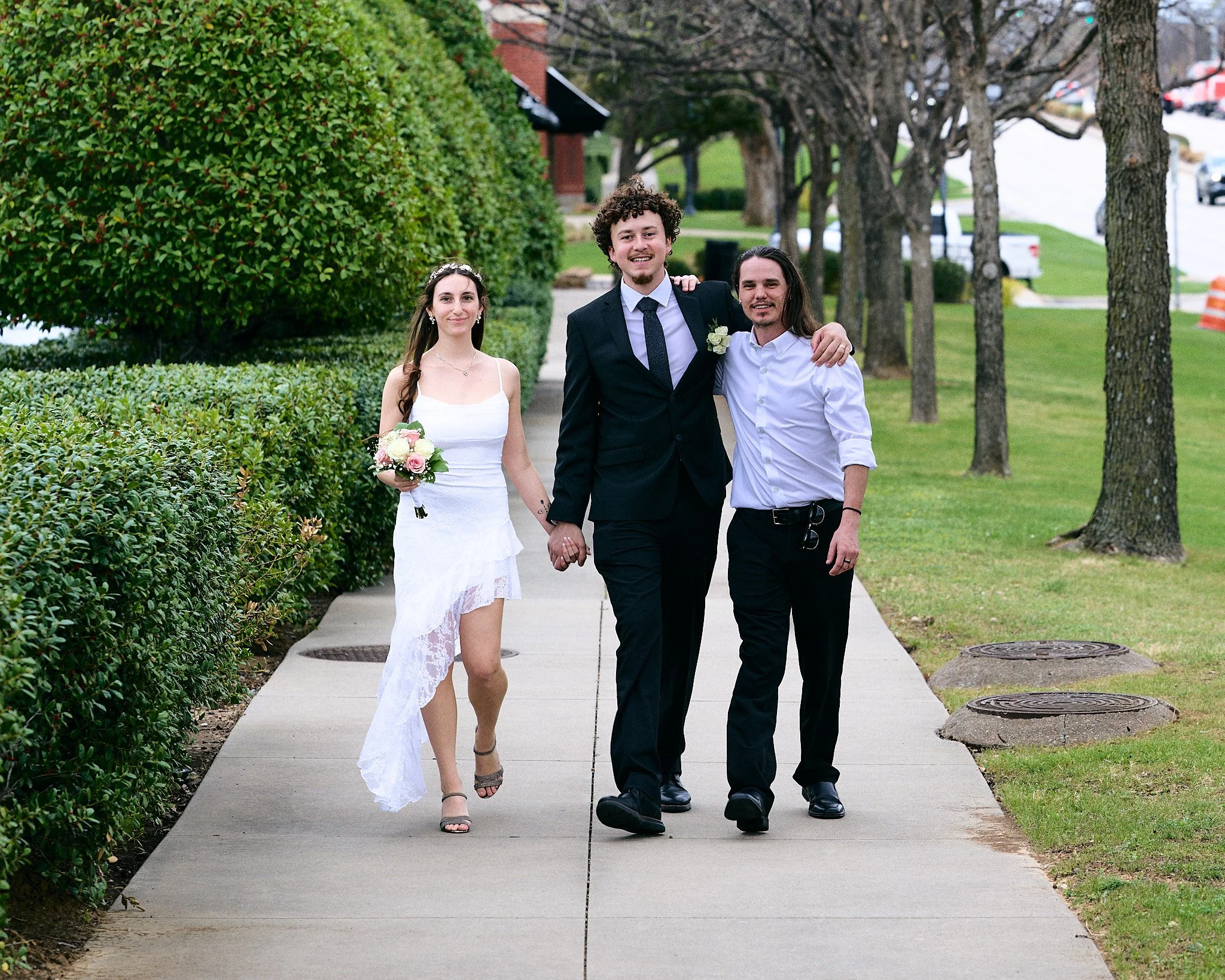 Three people walking on a sidewalk, dressed in formal attire. The woman on the left wears a white dress and holds a bouquet, the man in the middle is in a black suit with a boutonniere, and the man on the right is in a white shirt. They are smiling a