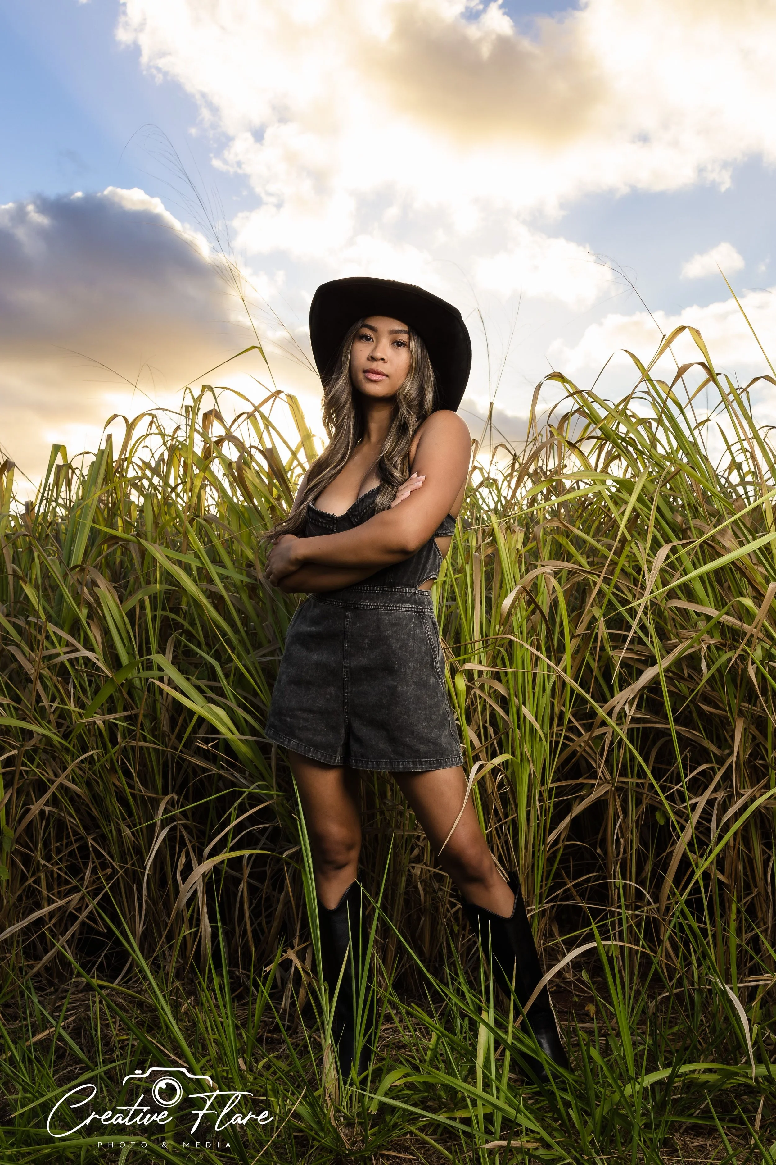 A woman standing in tall grass during sunset, wearing a black hat, black top, and black skirt, with her arms crossed.