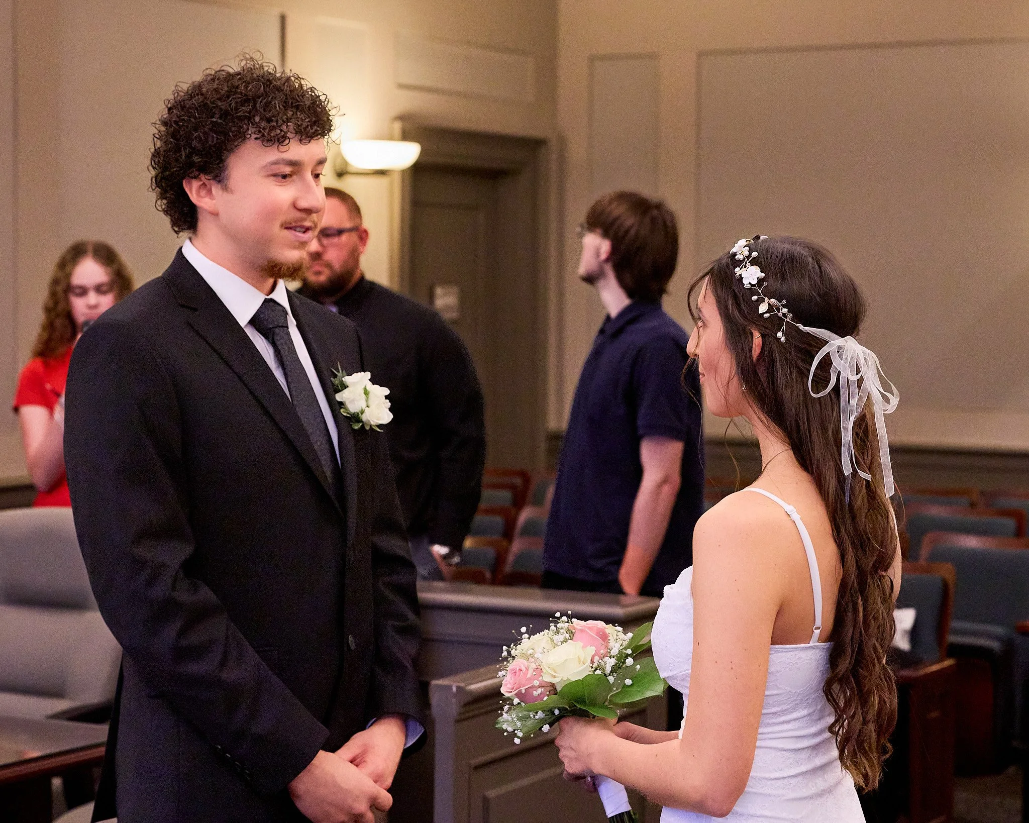 A groom in a black suit with a white shirt and black tie is standing in front of a bride holding a bouquet of white and pink flowers. The bride is wearing a white dress with spaghetti straps and a floral headpiece with white flowers and ribbons. They