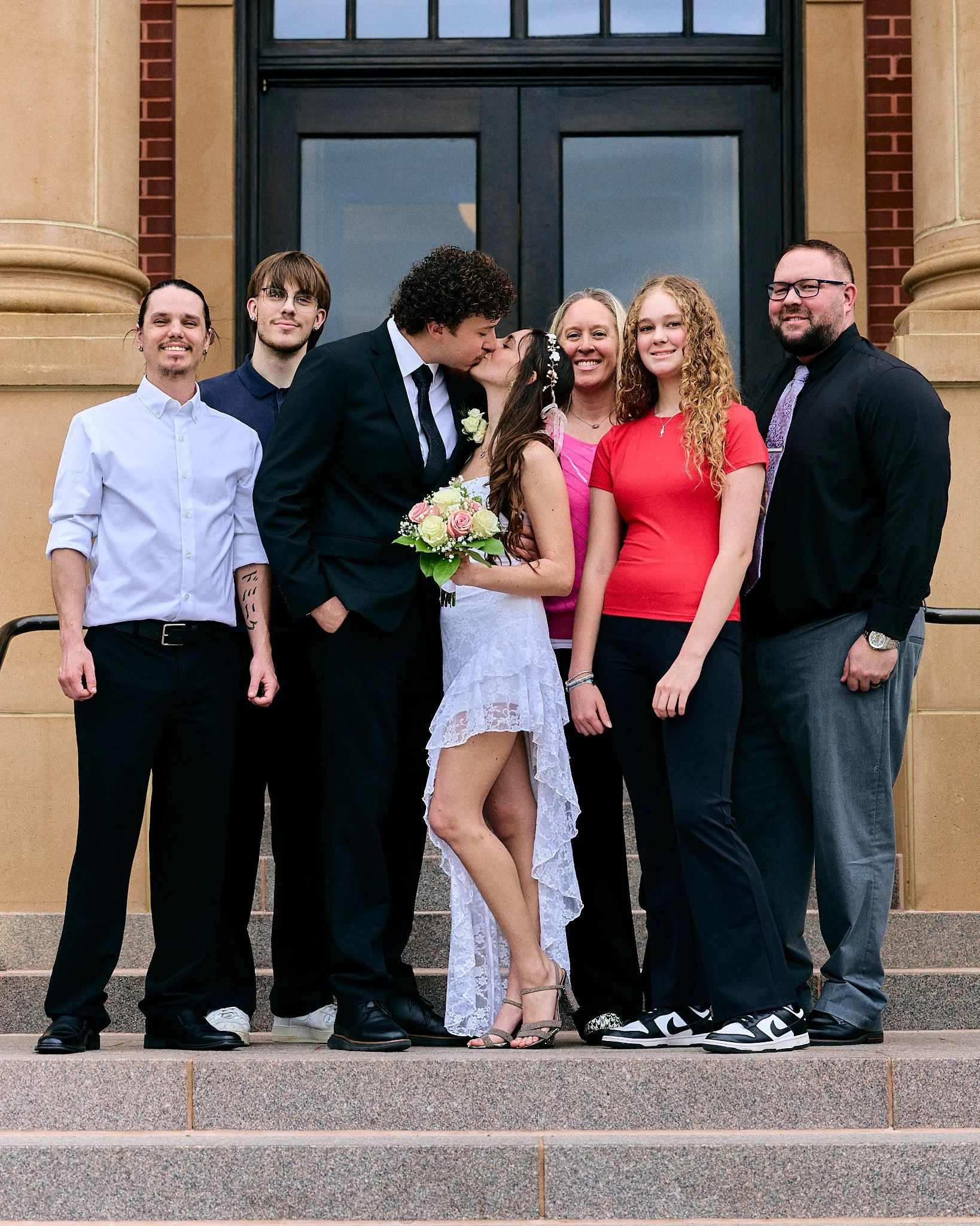 A group of seven people standing on outdoor steps in front of a building with large windows and brick and stone architectural details. A woman in a white dress holding a bouquet kisses a man in a black suit, and the others smile for the photo.