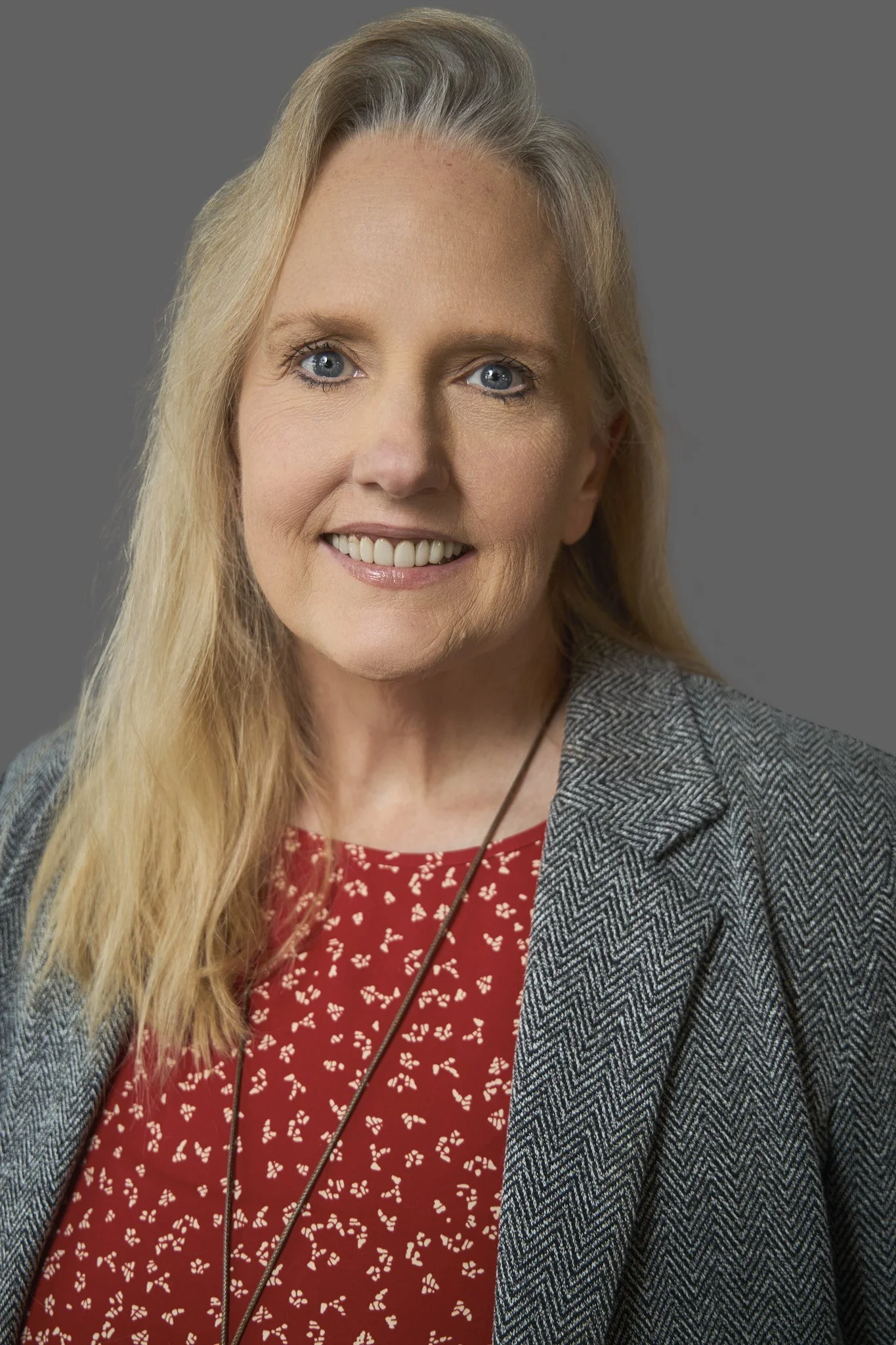 A middle-aged woman with long blonde hair, blue eyes, and a friendly smile, wearing a red blouse with white patterns and a gray herringbone blazer, standing against a gray background.