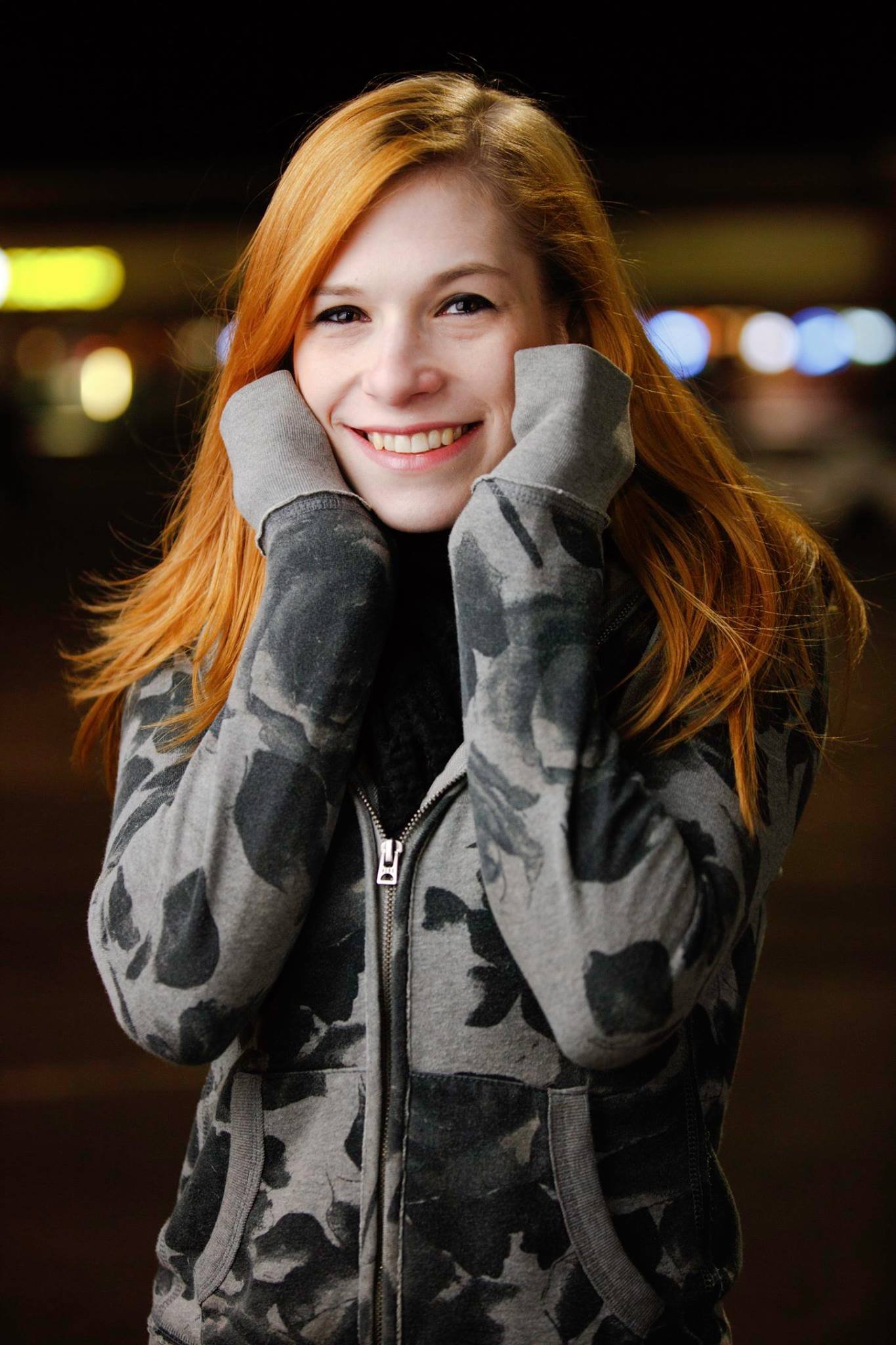 A young woman with long red hair smiling and holding her face with her hands in a camouflage hoodie, standing outdoors at night with blurred lights in the background.