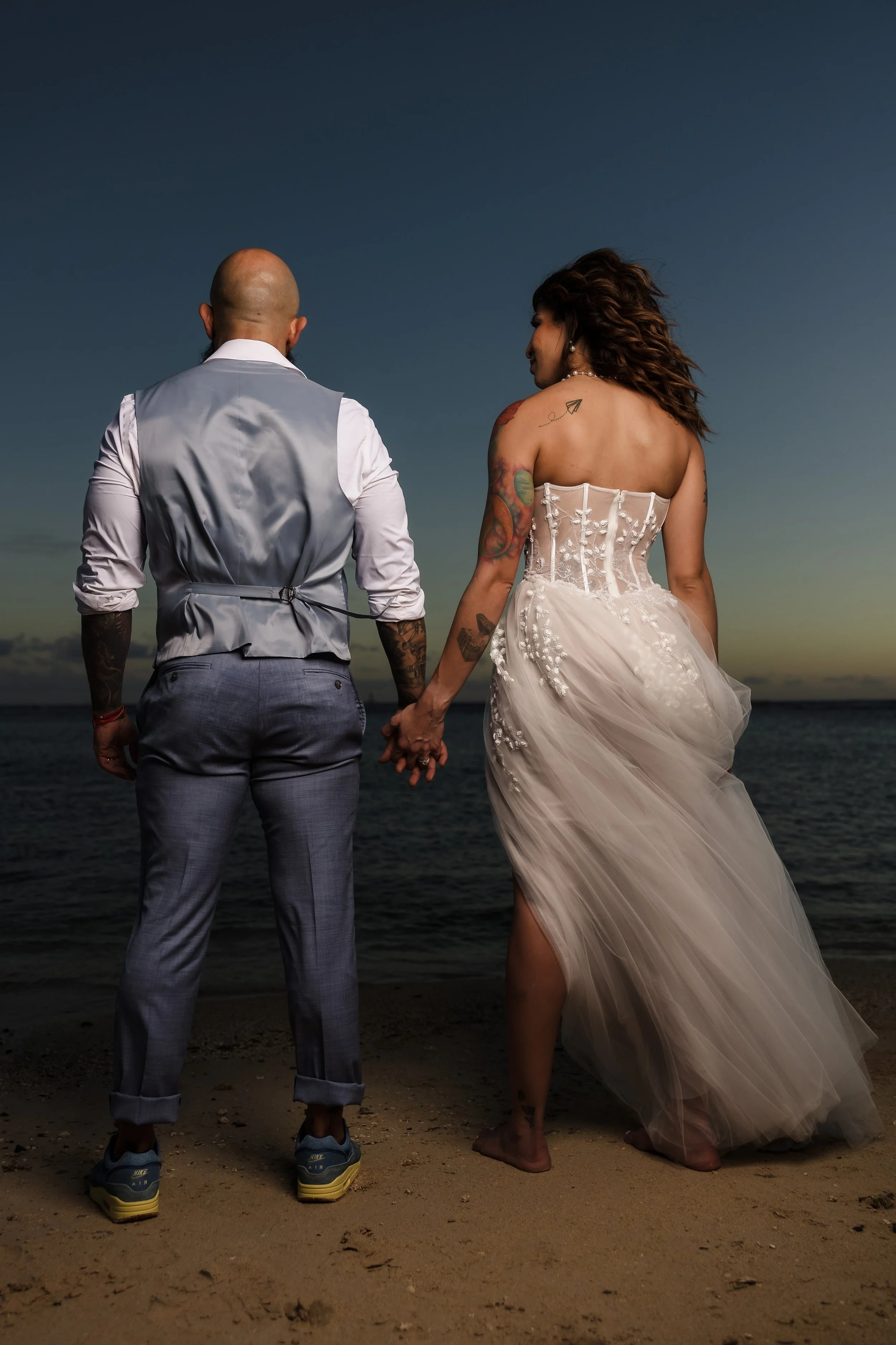 A couple dressed in wedding attire, holding hands, standing on a beach at sunset, facing the ocean.