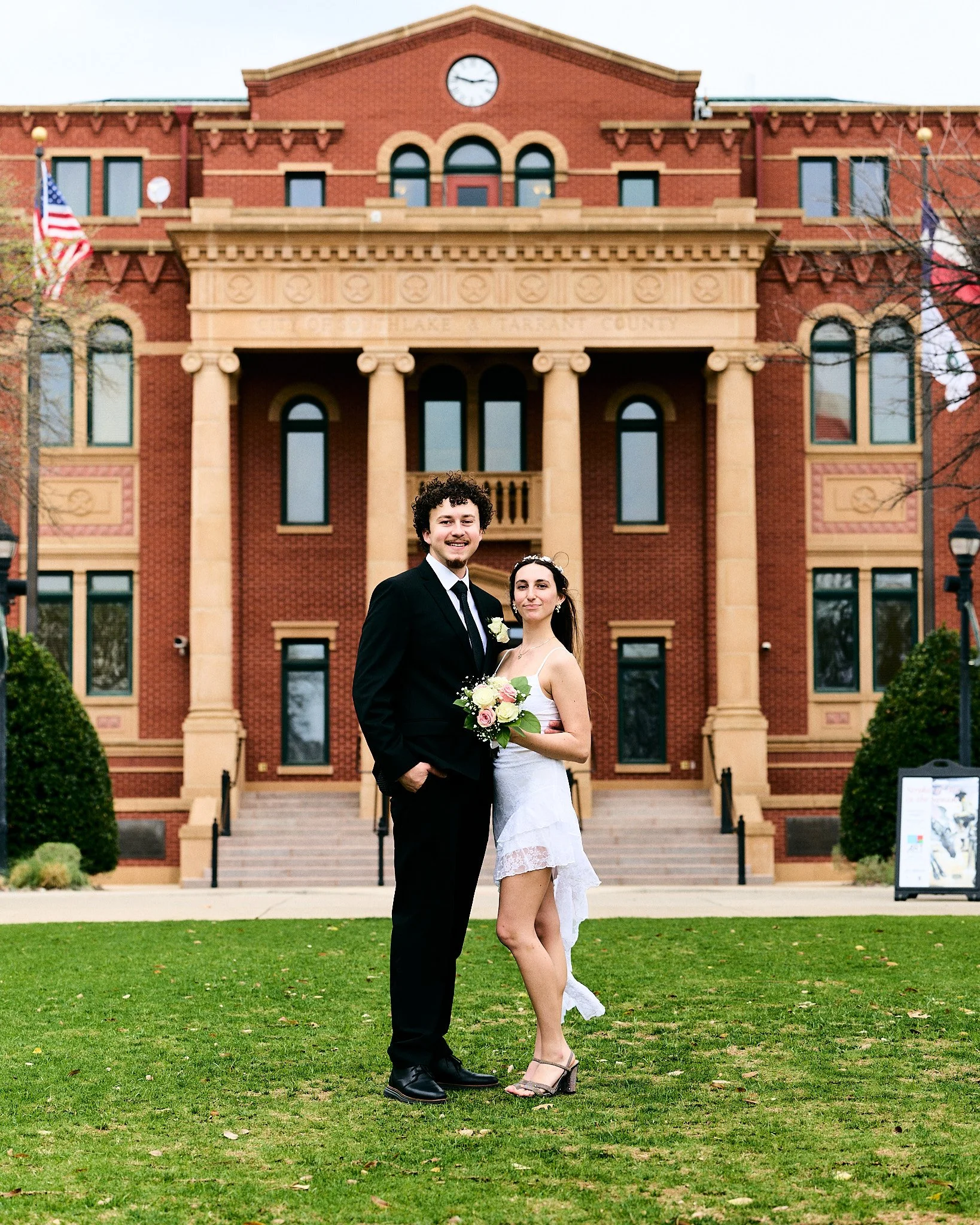 A bride and groom standing together, smiling, in front of a historic brick courthouse building, with American flags and steps leading up to the entrance.