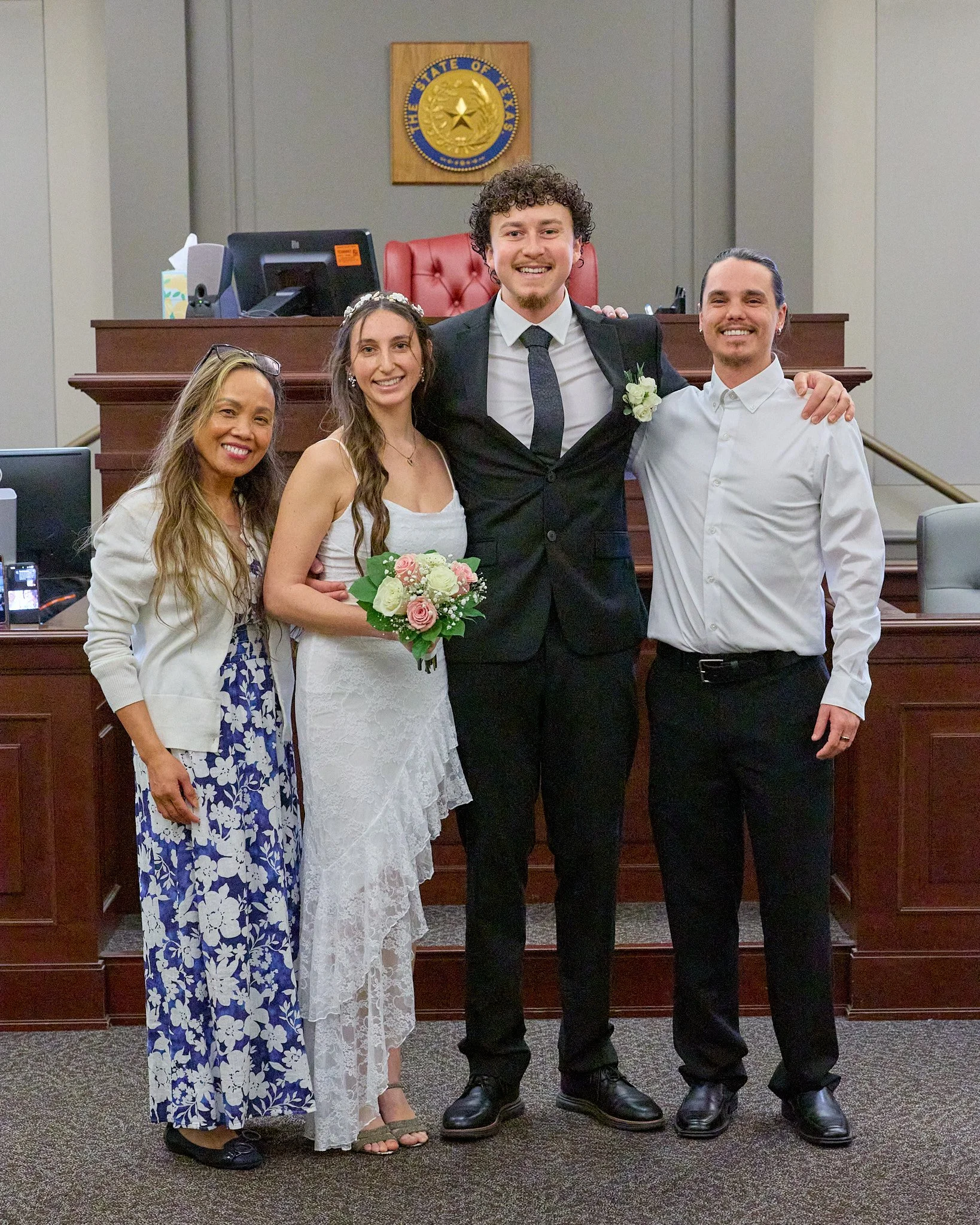 Four people, two women and two men, standing in a courtroom, smiling and posing for a photo. The woman in the middle is wearing a white dress and holding a bouquet, and the man in the center has his arm around her. The background features a judge's b