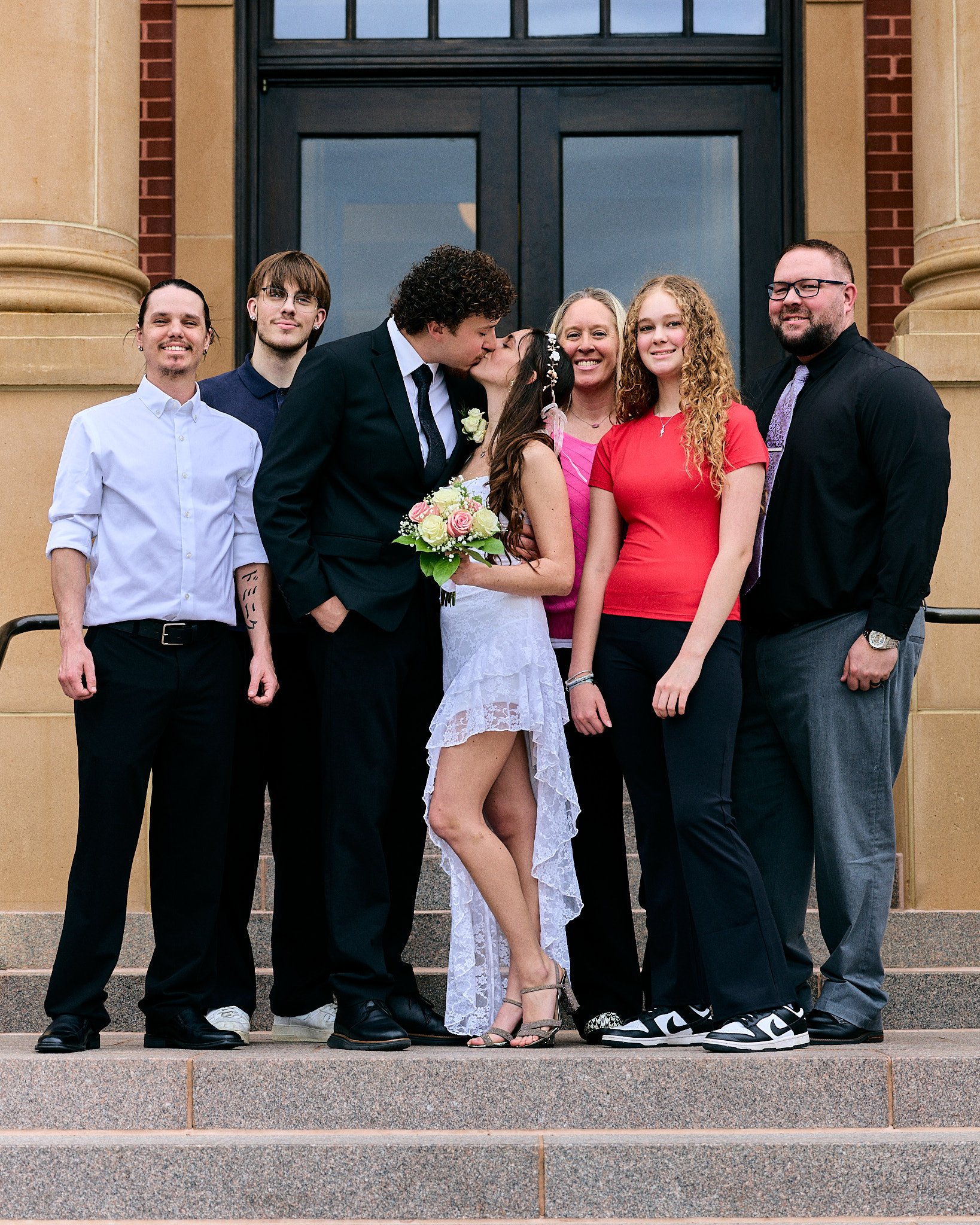 A group of seven people standing on steps in front of a building with large glass doors. Two people in the middle are kissing, one man and one woman. The woman is holding a bouquet of flowers. The group is smiling and dressed semi-formally, with four