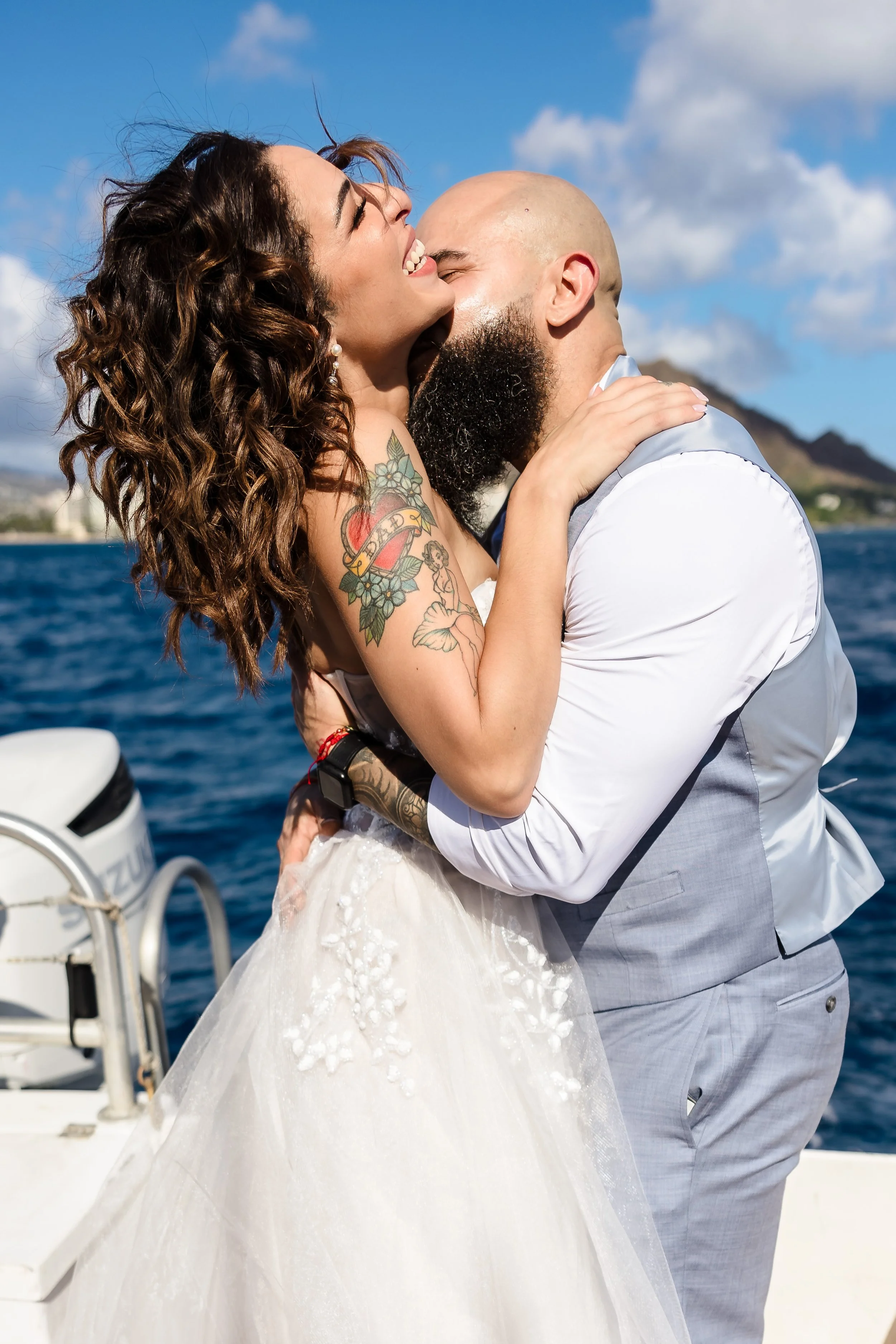 A newlywed couple sharing a joyful embrace on a boat with water and mountains in the background, under a partly cloudy sky.