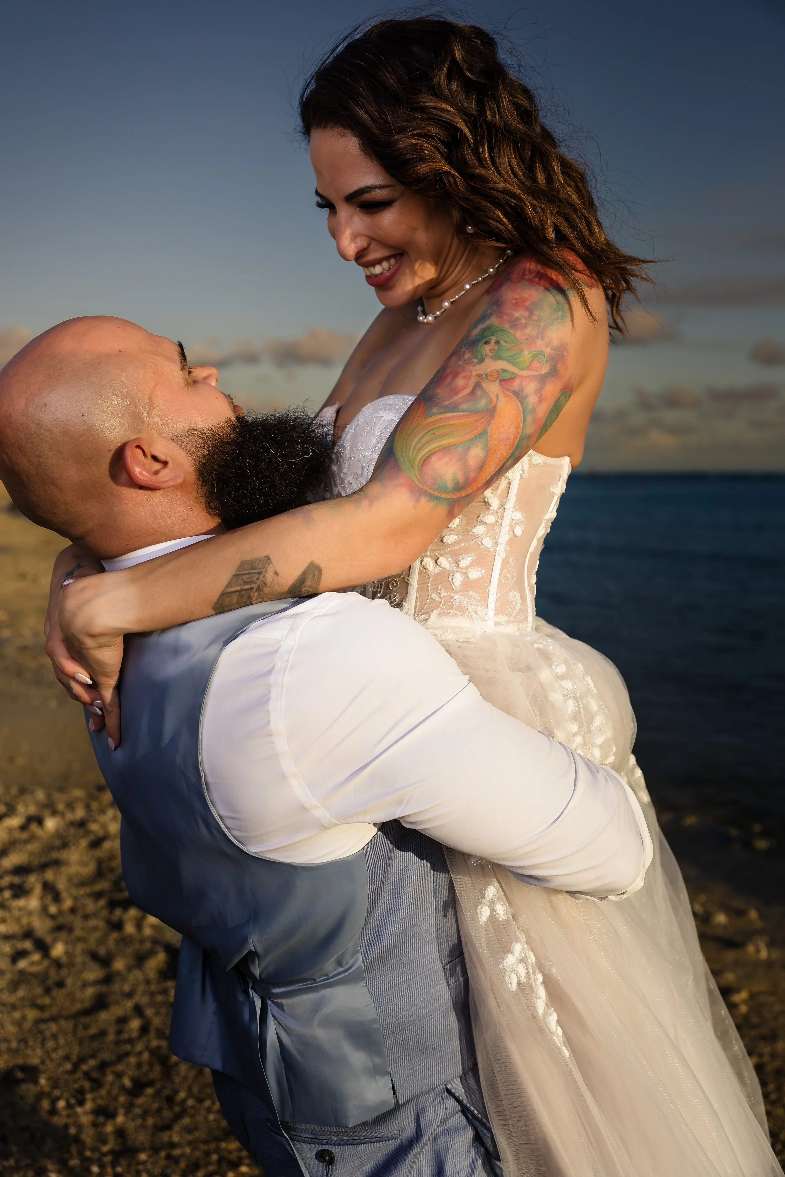 A happy couple at the beach, with the woman in a wedding dress and the man in a suit, embracing and smiling at each other during sunset.