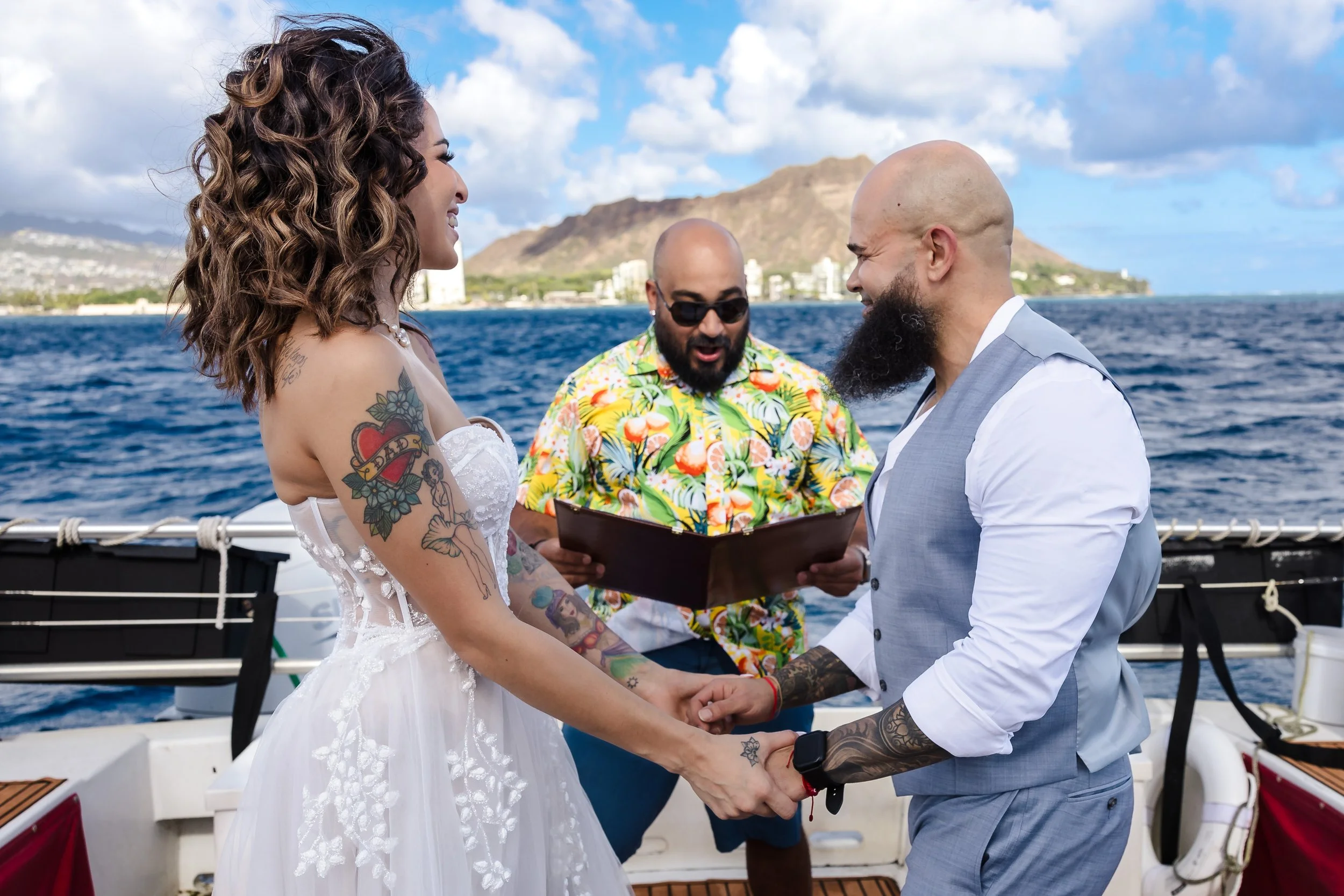 A woman and a man holding hands during a wedding ceremony on a boat, with a bald officiant in sunglasses and a Hawaiian shirt officiating, with mountains and ocean in the background.