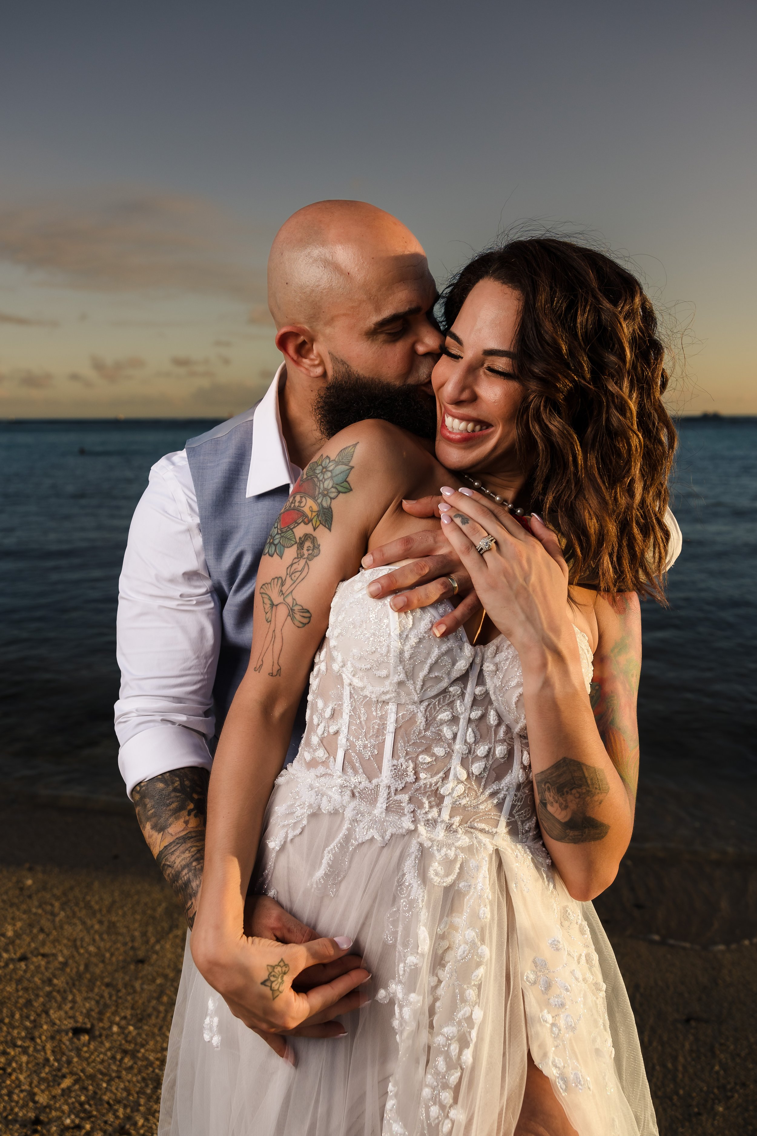 A newlywed couple embraces at the beach during sunset. The groom is kissing the bride on the cheek, both smiling. The bride has long wavy hair and visible tattoos on her shoulder and arm, and wears a white lace wedding dress. The groom is bald, has a