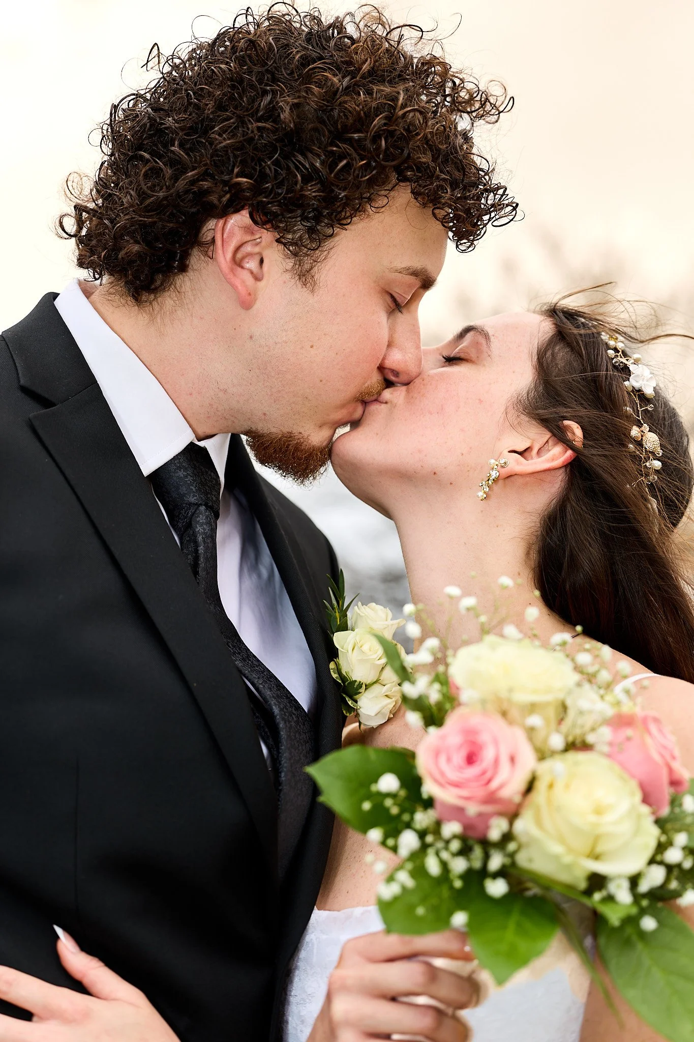 A close-up of a bride and groom kissing at their wedding, with the bride holding a bouquet of pink and white roses and greenery.