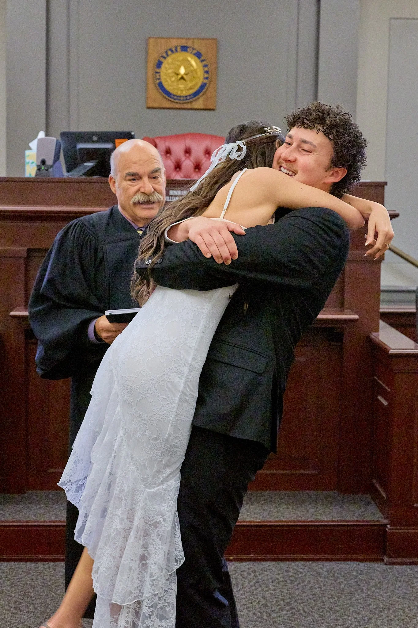 A groom and bride share a hug in a courthouse, with a judge observing. The judge is standing beside them, holding a small book, wearing a black robe. The background features a courthouse seal on the wall, a wooden bench, and a computer on a desk.