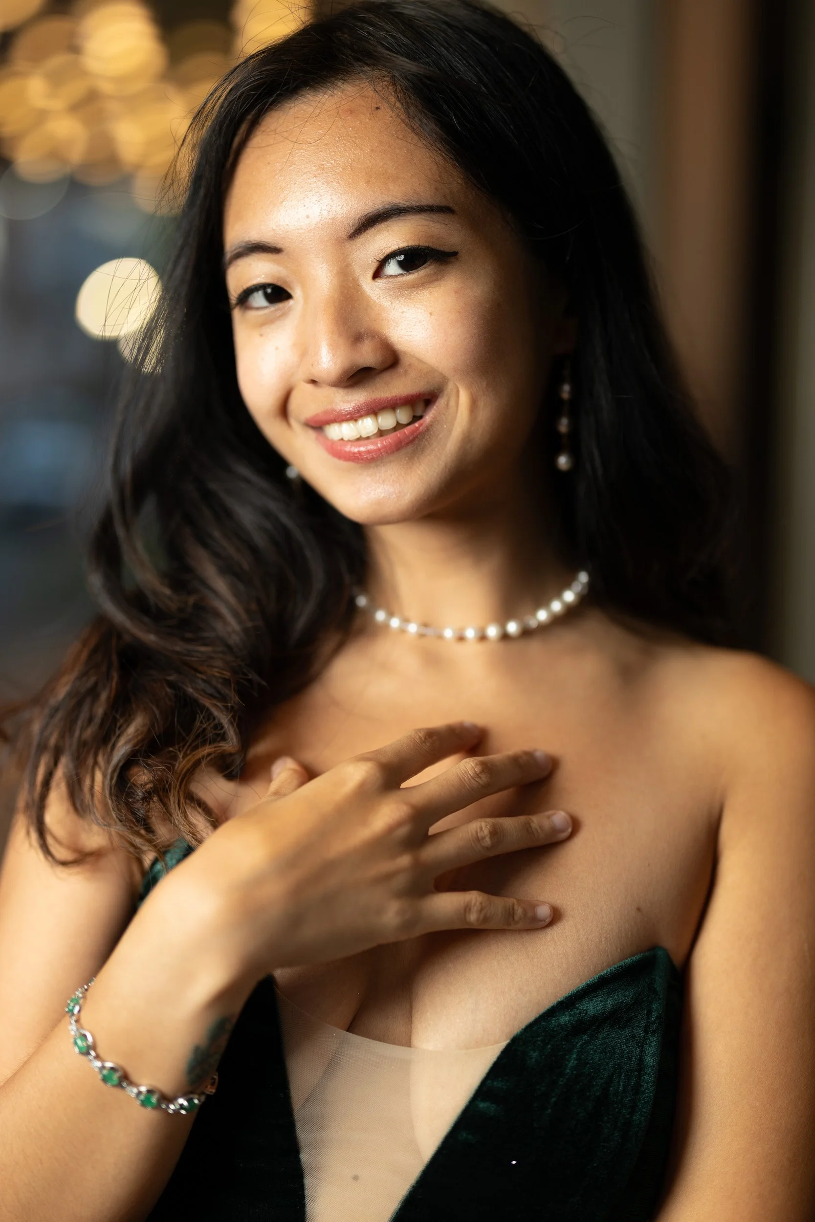 A woman smiling, holding her hand on her chest, wearing jewelry including pearl earrings, a pearl necklace, and a bracelet, dressed in a dark green and black strapless dress with a beige inner lining.