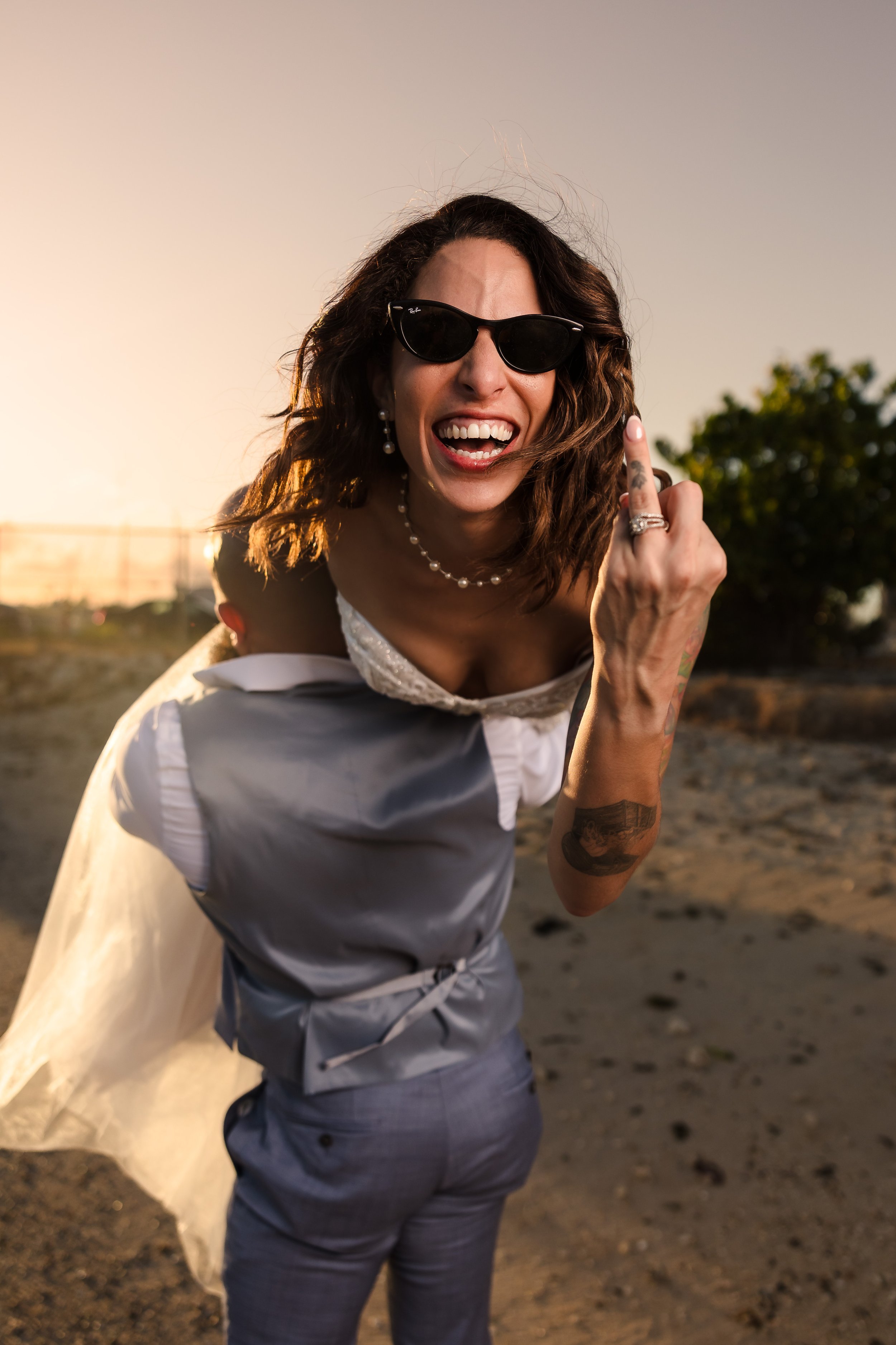 Woman with curly hair wearing sunglasses, earrings, and a necklace, flipping off the camera with her middle finger, outdoors during sunset.