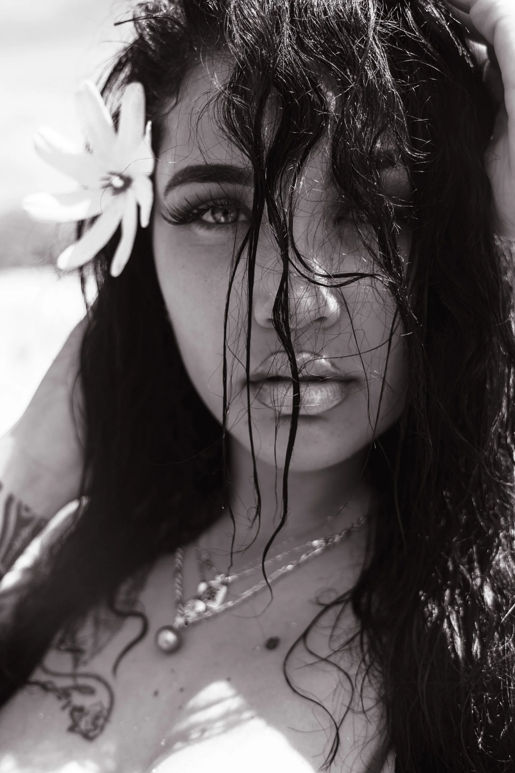 Black and white close-up photo of a woman with wet and curly hair, holding a flower near her face, with tattoos on her arm and a zipper on her outfit.