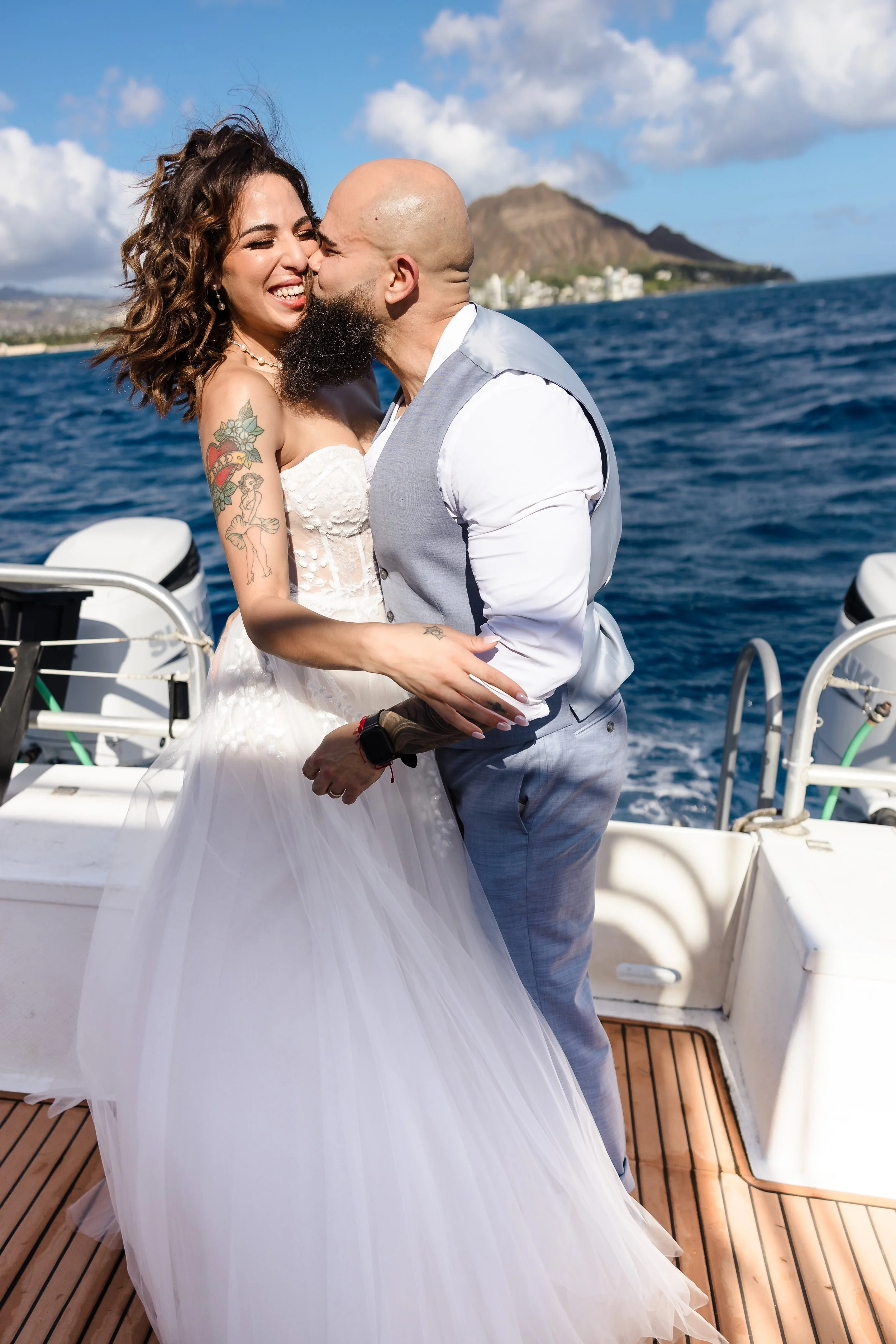 A couple on a boat, with the man kissing the woman's cheek as she smiles and laughs, in front of a background of water, mountains, and a city skyline.