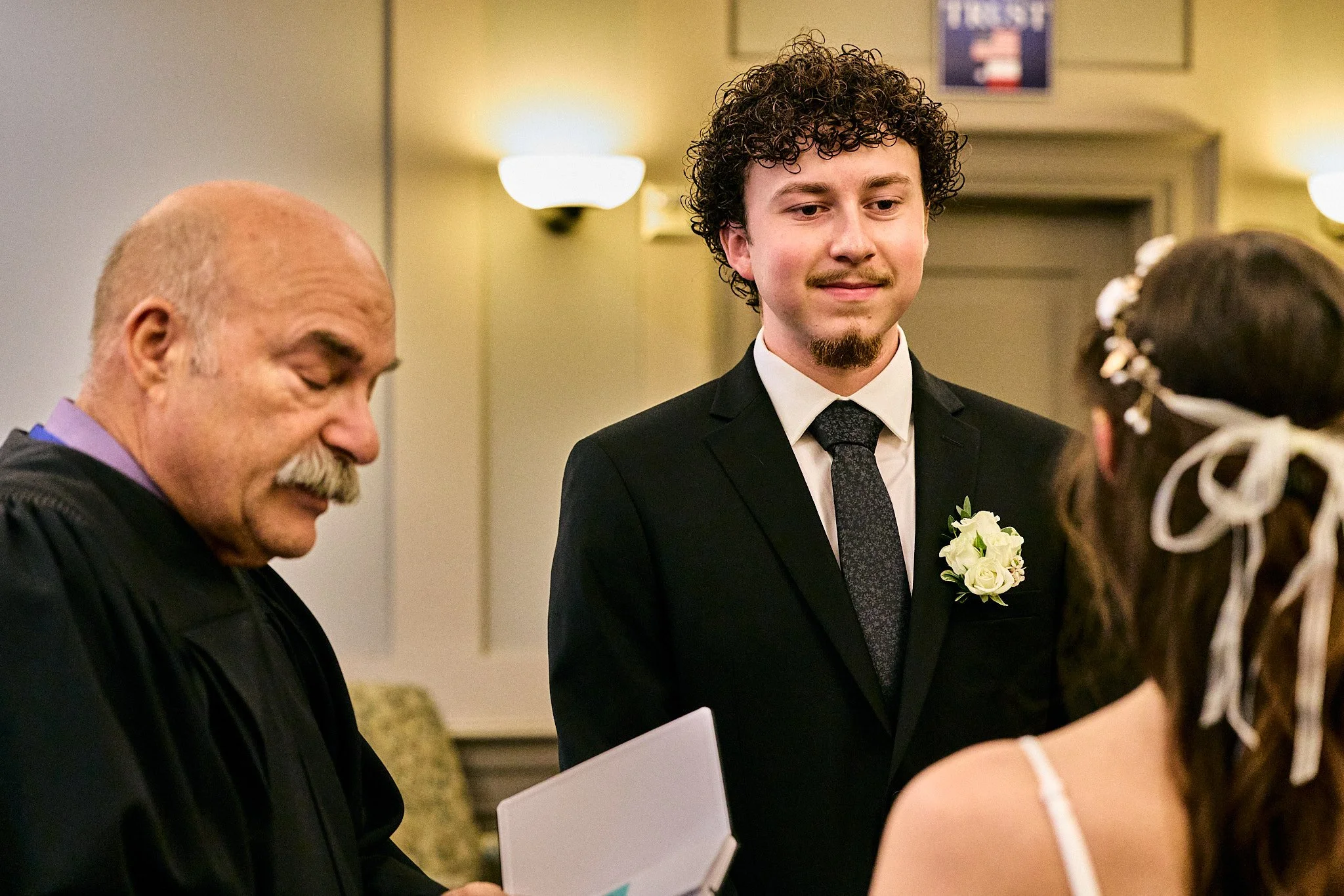 A groom in a black suit with a boutonniere on his lapel stands during a wedding ceremony, with a woman in white attire, likely the bride, in the foreground. An officiant in purple robes is seen on the left, and a person with dark hair and white flowe