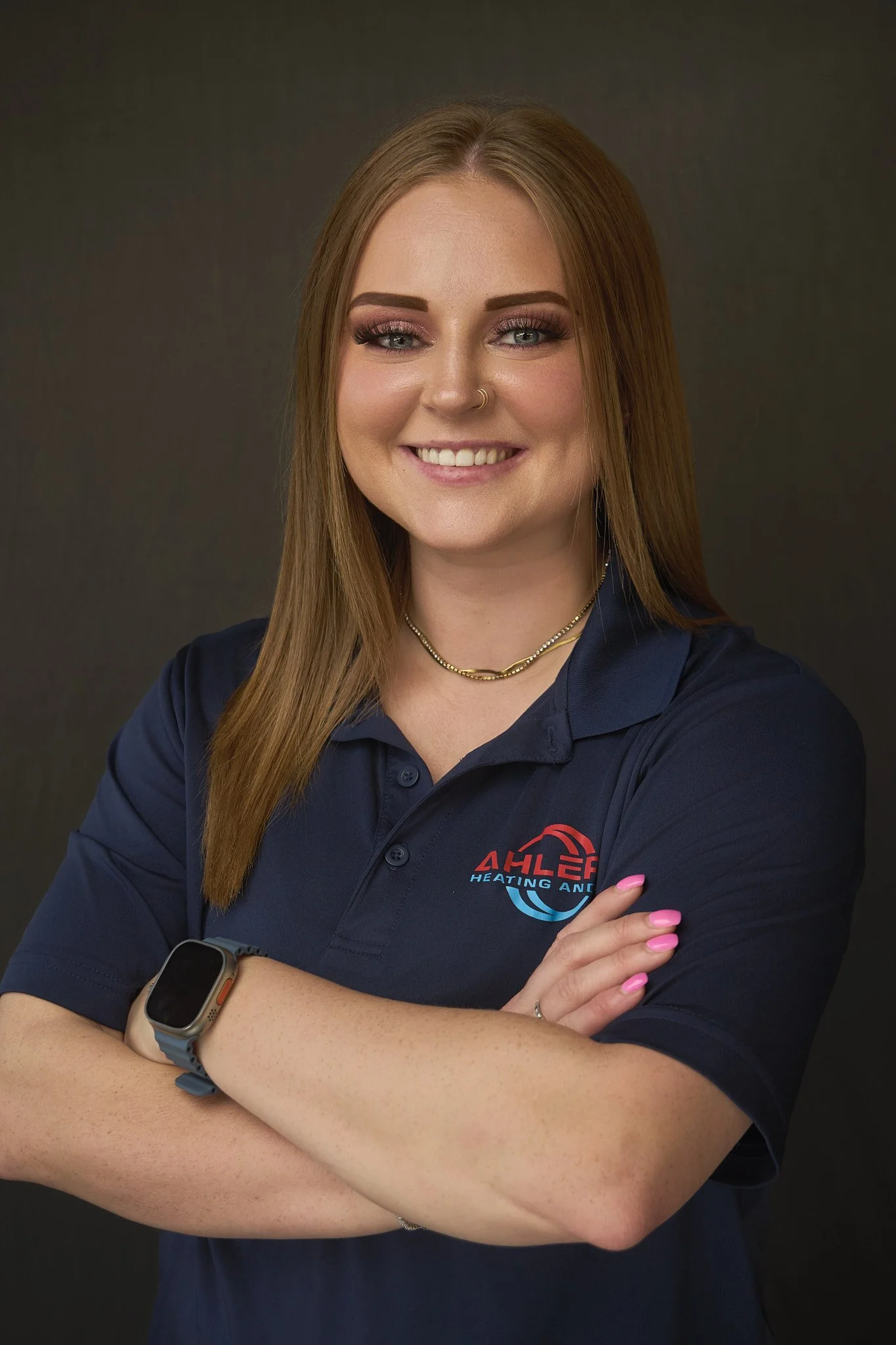 Close-up portrait of a smiling woman with long straight red hair, blue eyes, wearing a navy blue polo shirt with a company logo reading 'AHLEF Heating and AC,' a gold necklace, and a smartwatch, standing against a dark background.