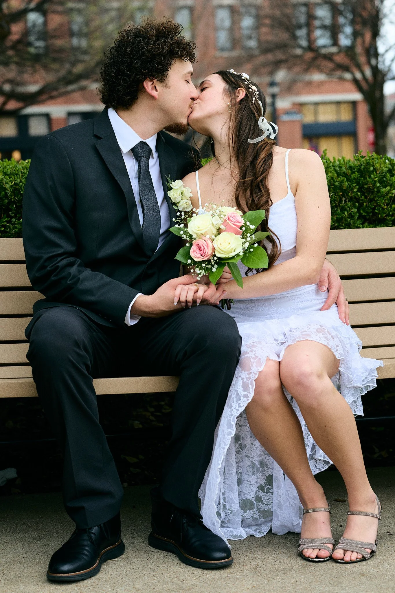 A couple in wedding attire sharing a kiss on a park bench, with the bride holding a bouquet of flowers, and trees in the background.