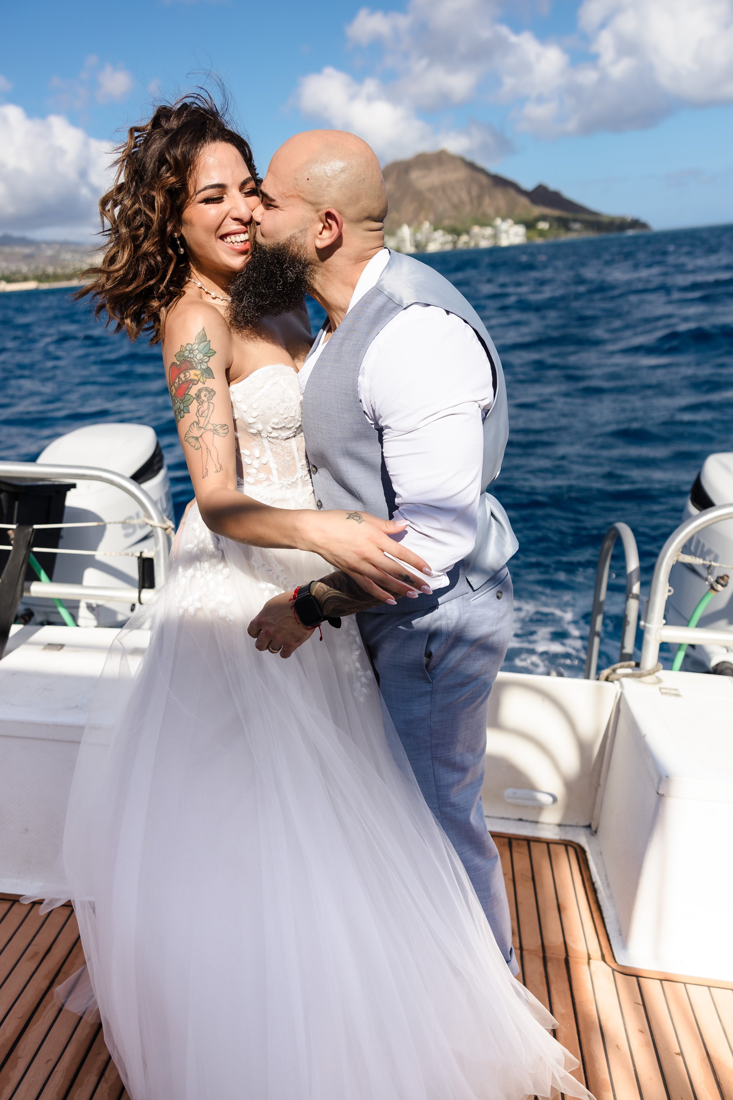 A couple on a boat, embracing and kissing with a mountain and city in the background, with blue sky and clouds above.