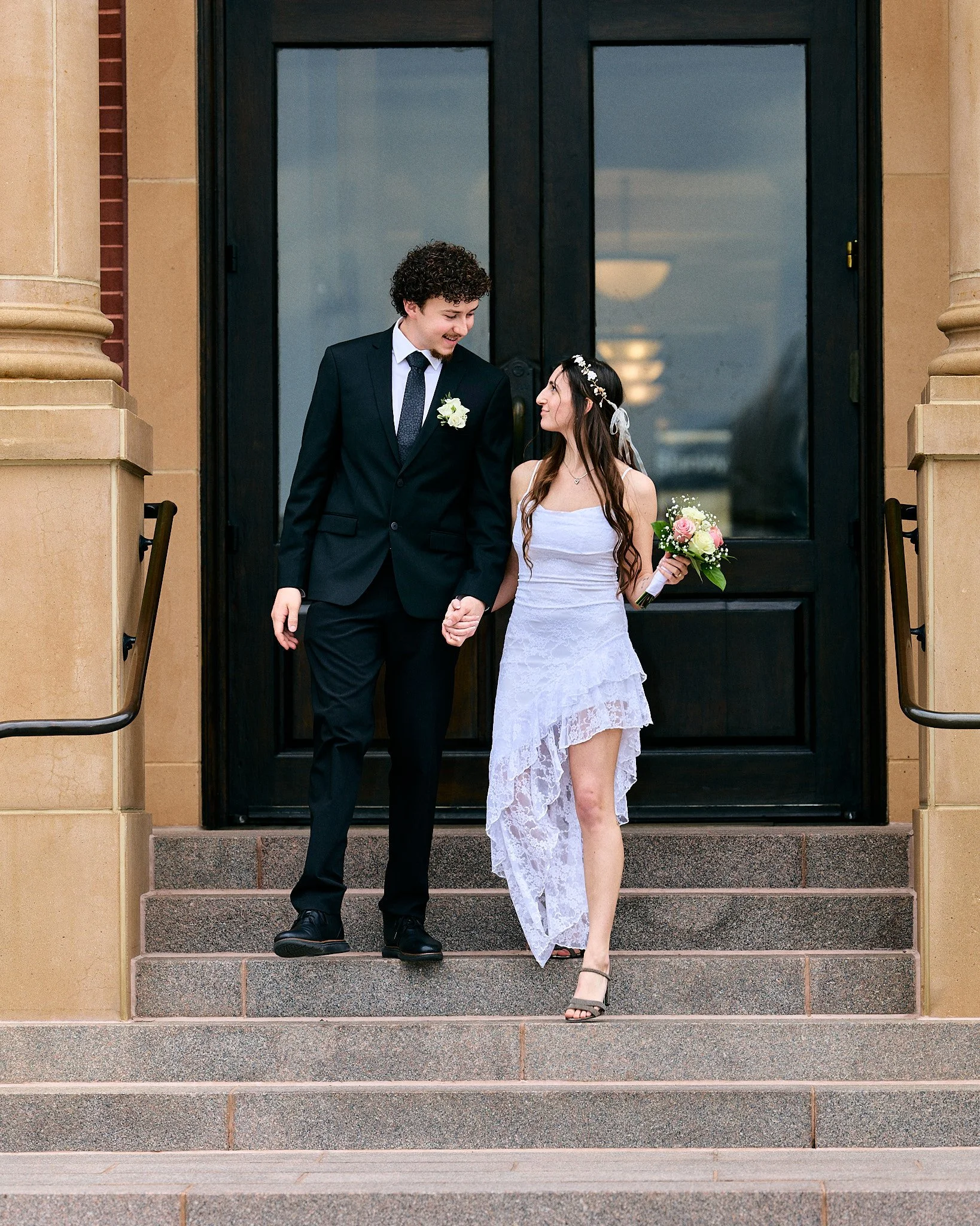 A bride and groom standing on the steps outside a building, holding hands and smiling at each other. The groom is wearing a black suit and tie with a white shirt and a boutonniere. The bride is wearing a white dress with lace details, a floral headba