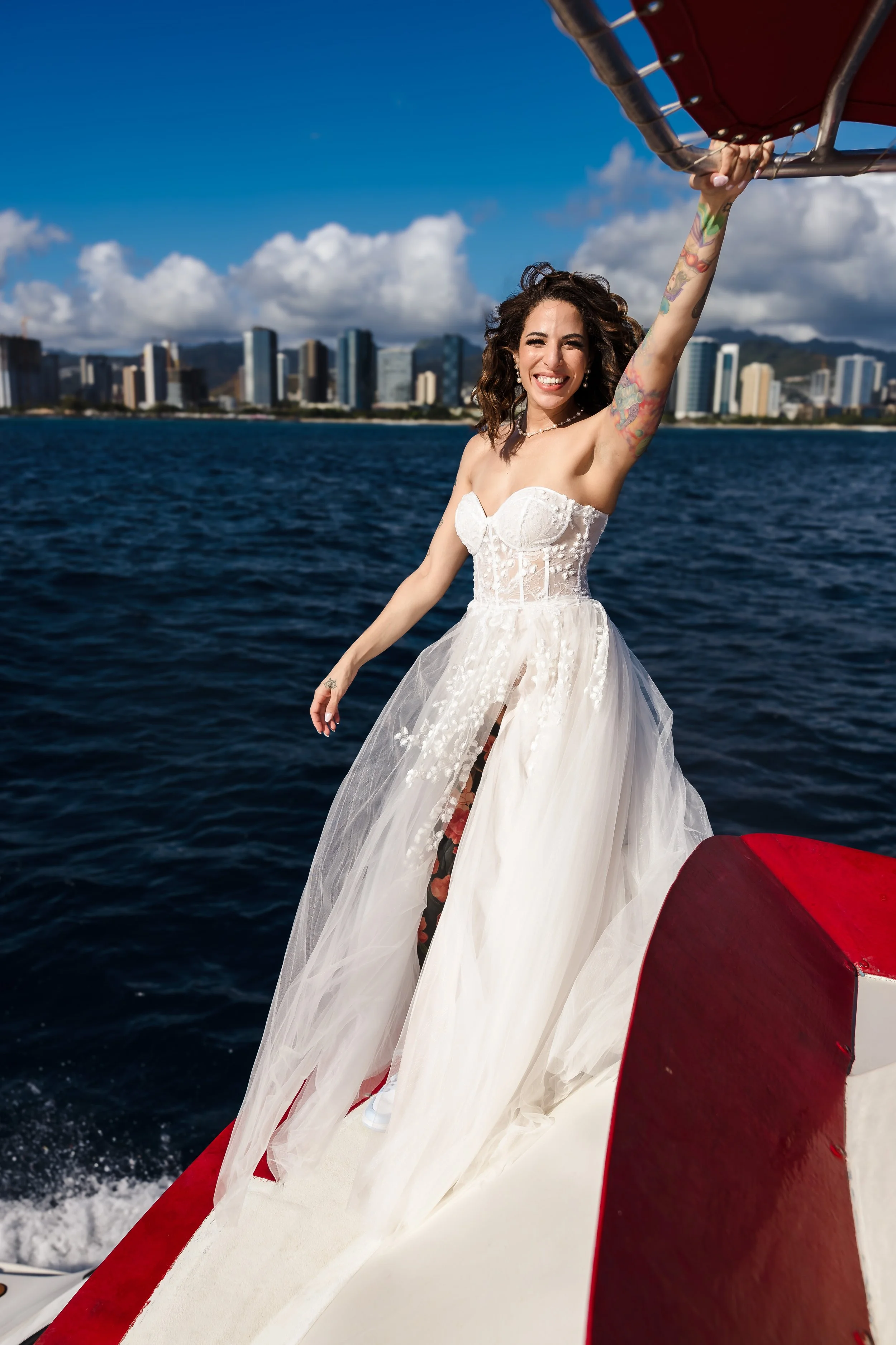 Woman in a wedding dress smiling and waving from a boat on the water with a city skyline and clouds in the background.