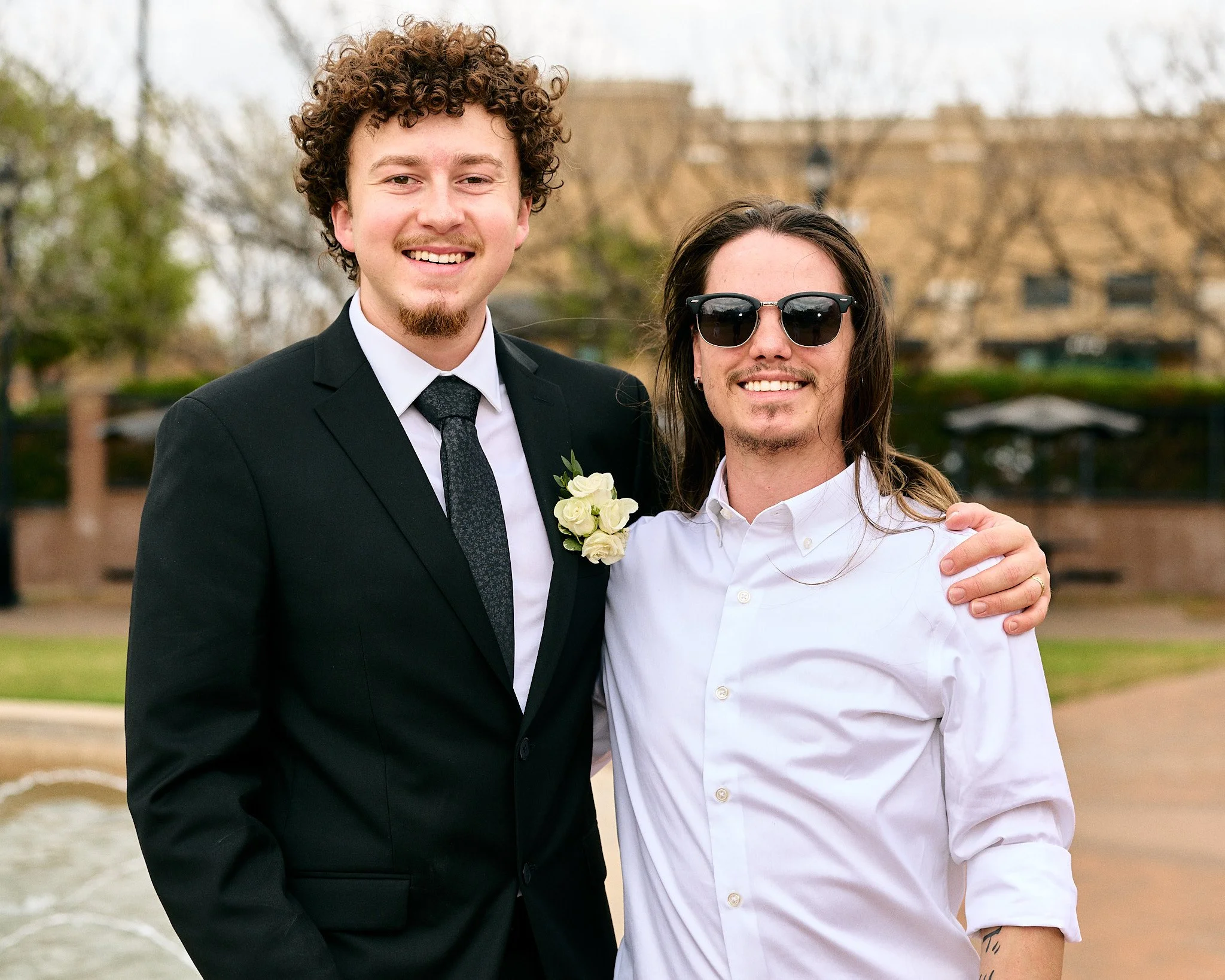 Two men standing outdoors near water, smiling and with arms around each other. One in a black suit and the other in a white shirt, wearing sunglasses.